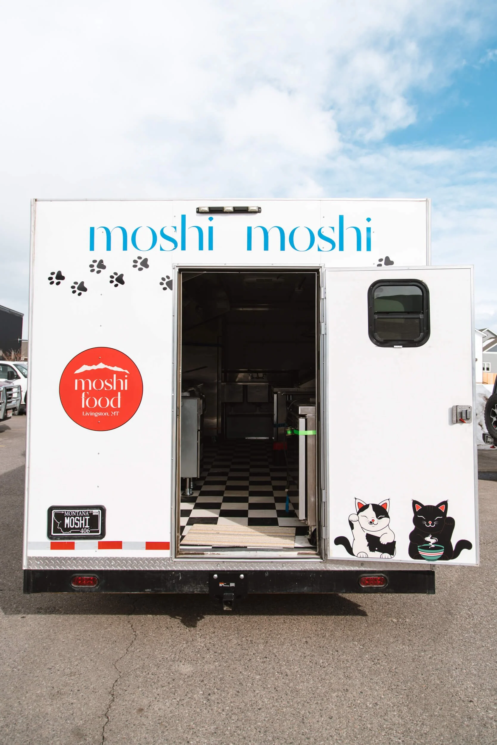 Back of Moshi Food trailer in Livingston, MT, showcasing “moshi moshi” wordmark, playful cat illustrations, and paw print trail leading to an open food truck interior.