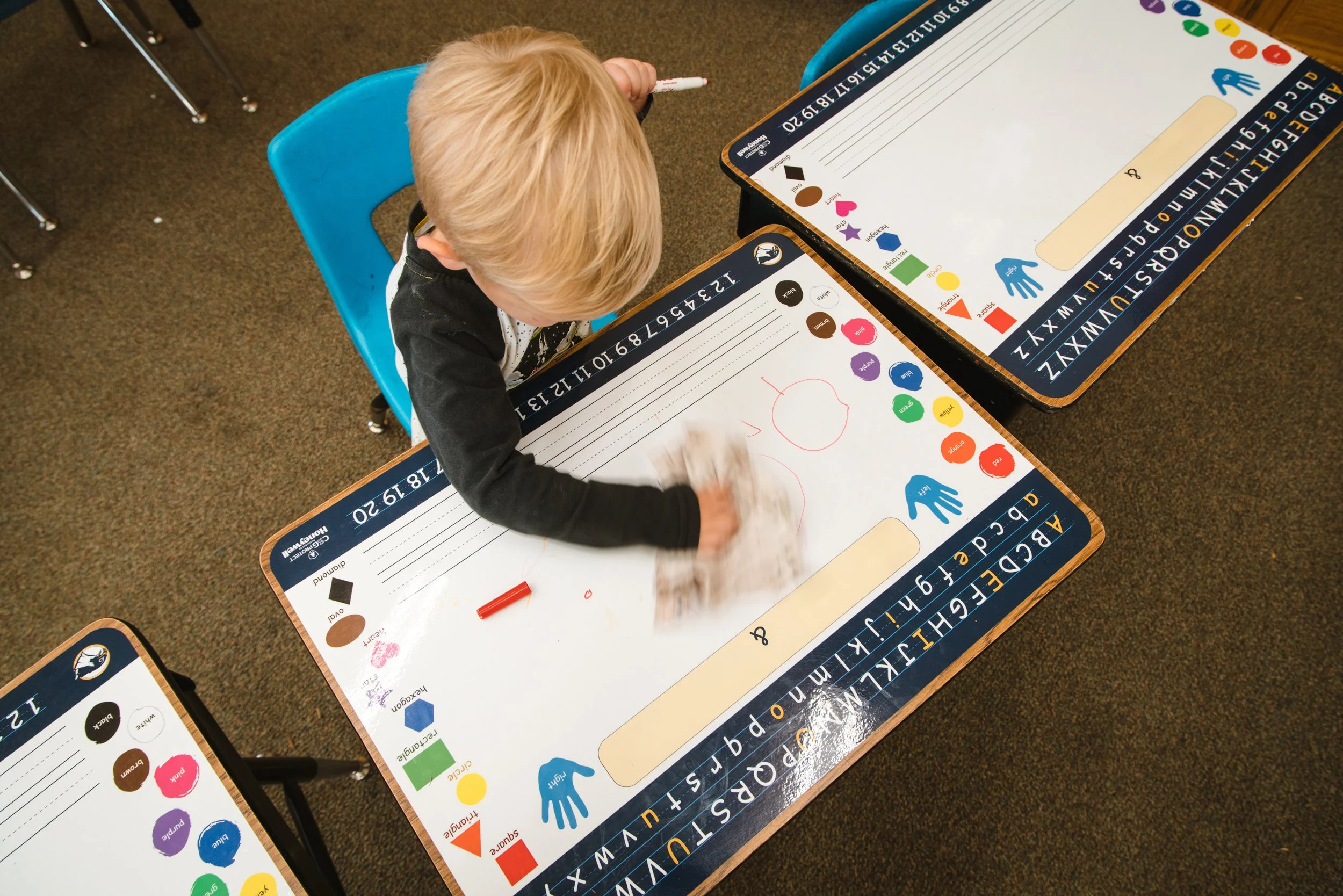 Overhead view of a young student wiping a printed dry erase desk wrap with a cloth.