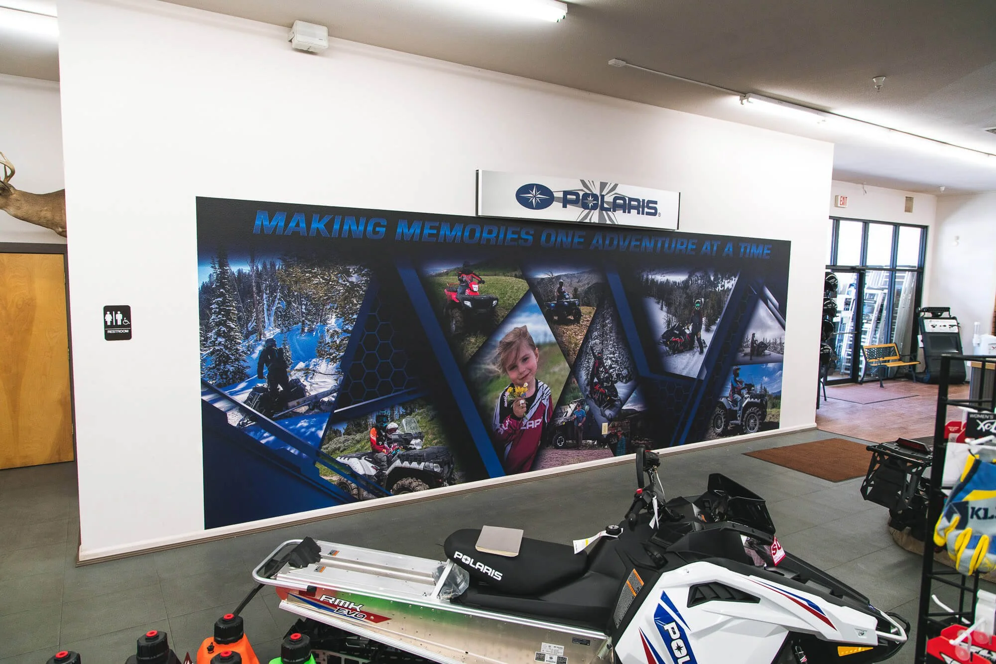 Vibrant showroom wall wrap for Polaris showcasing dynamic outdoor scenes of snowmobiling and off-roading with the slogan “Making Memories One Adventure at a Time,” adding energy and brand identity to the retail space in Bozeman, MT.