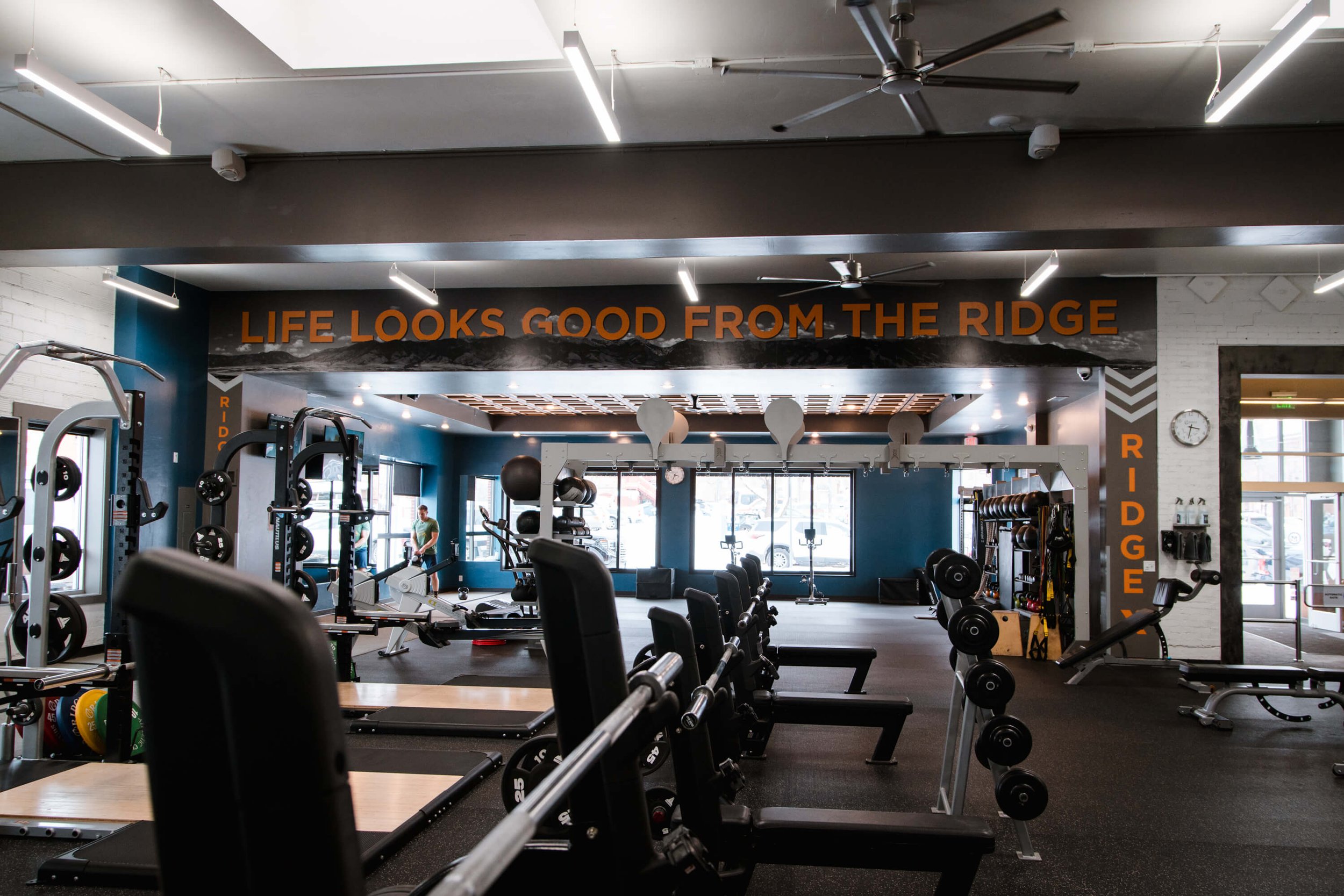 Panoramic view of a modern gym interior featuring a wall wrap above the weightlifting area that reads “LIFE LOOKS GOOD FROM THE RIDGE” in bold orange lettering with mountain imagery in the background, showcasing motivational fitness branding. Locatio