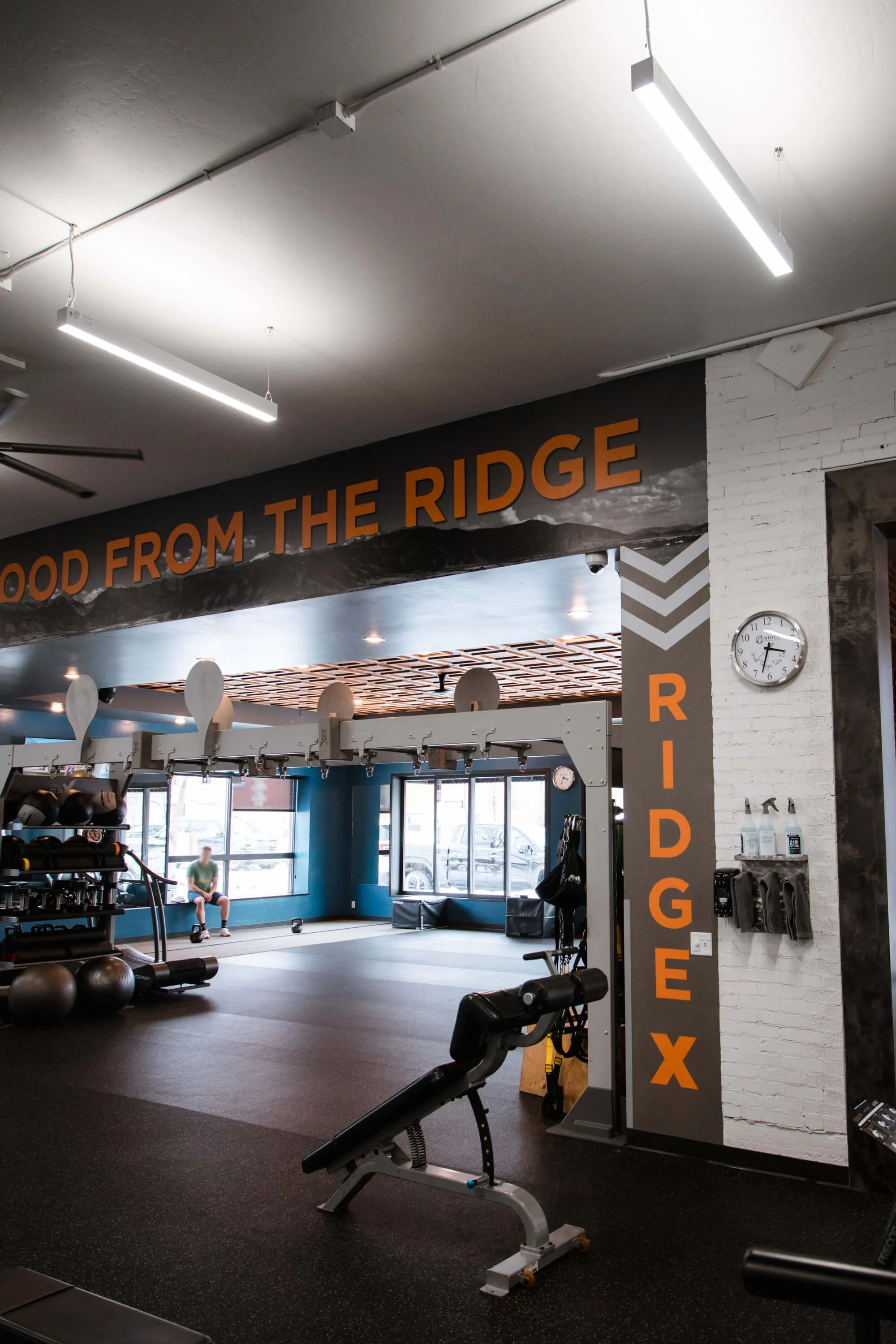 Close-up of a fitness studio wall wrap featuring the word “RIDGEX” vertically aligned and “LIFE LOOKS GOOD FROM THE RIDGE” horizontally, both in vibrant orange typography layered over a grayscale mountain graphic. Location: The Ridge, Bozeman, MT.