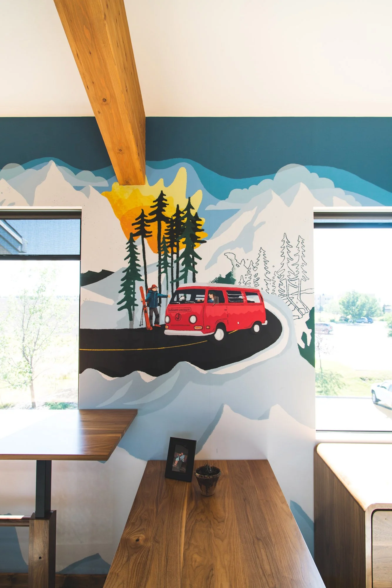 Colorful custom wall wrap featuring a red Volkswagen van, skier, pine trees, and a sunset against a mountainous backdrop, designed for an indoor office wall space at what was formerly Jelt Belt in Bozeman, MT.