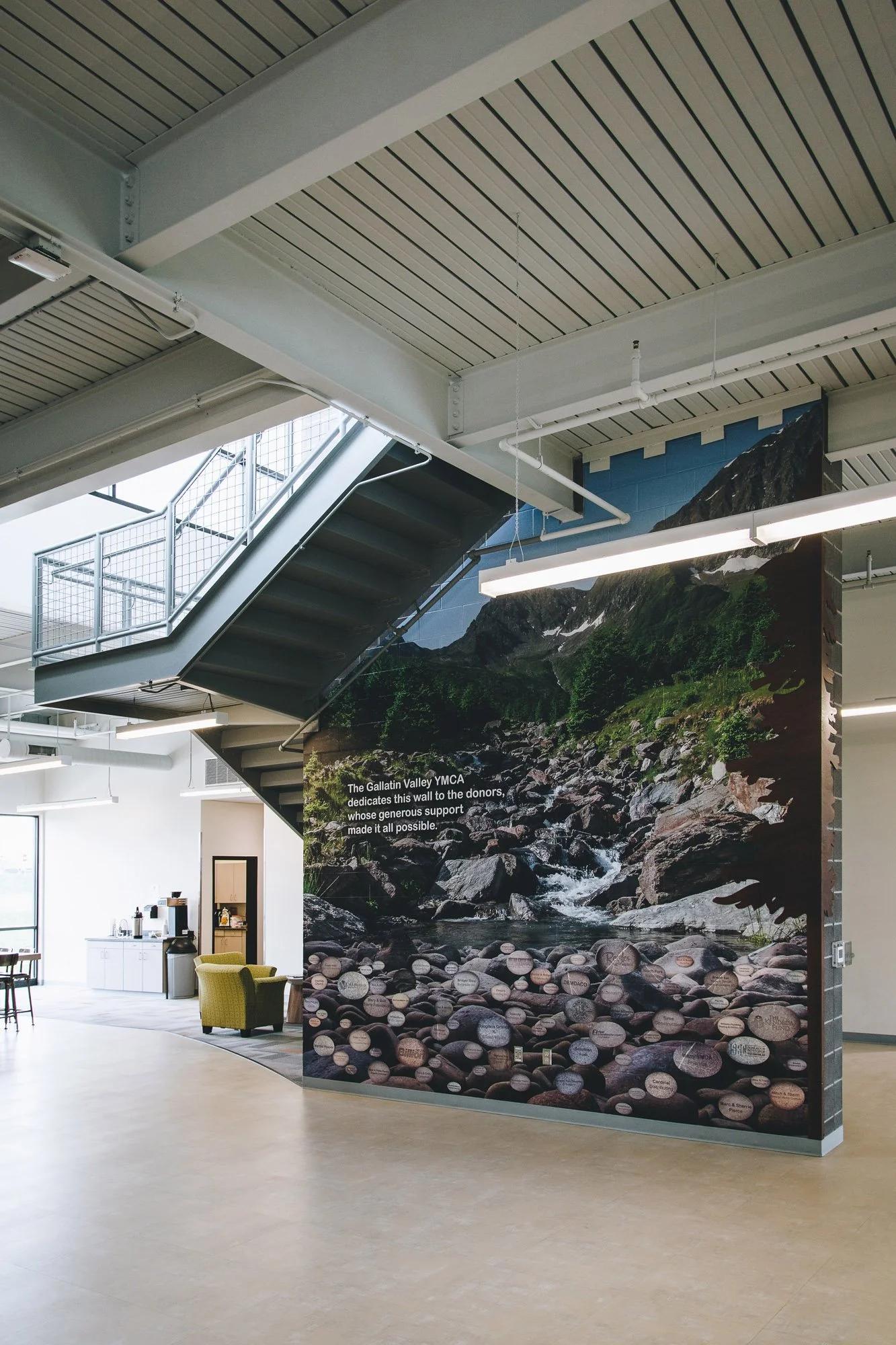 Large interior wall wrap featuring a scenic photograph of a forested stream with inspirational text, installed in a modern stairwell as part of a donor recognition display at the YMCA in Bozeman, MT.
