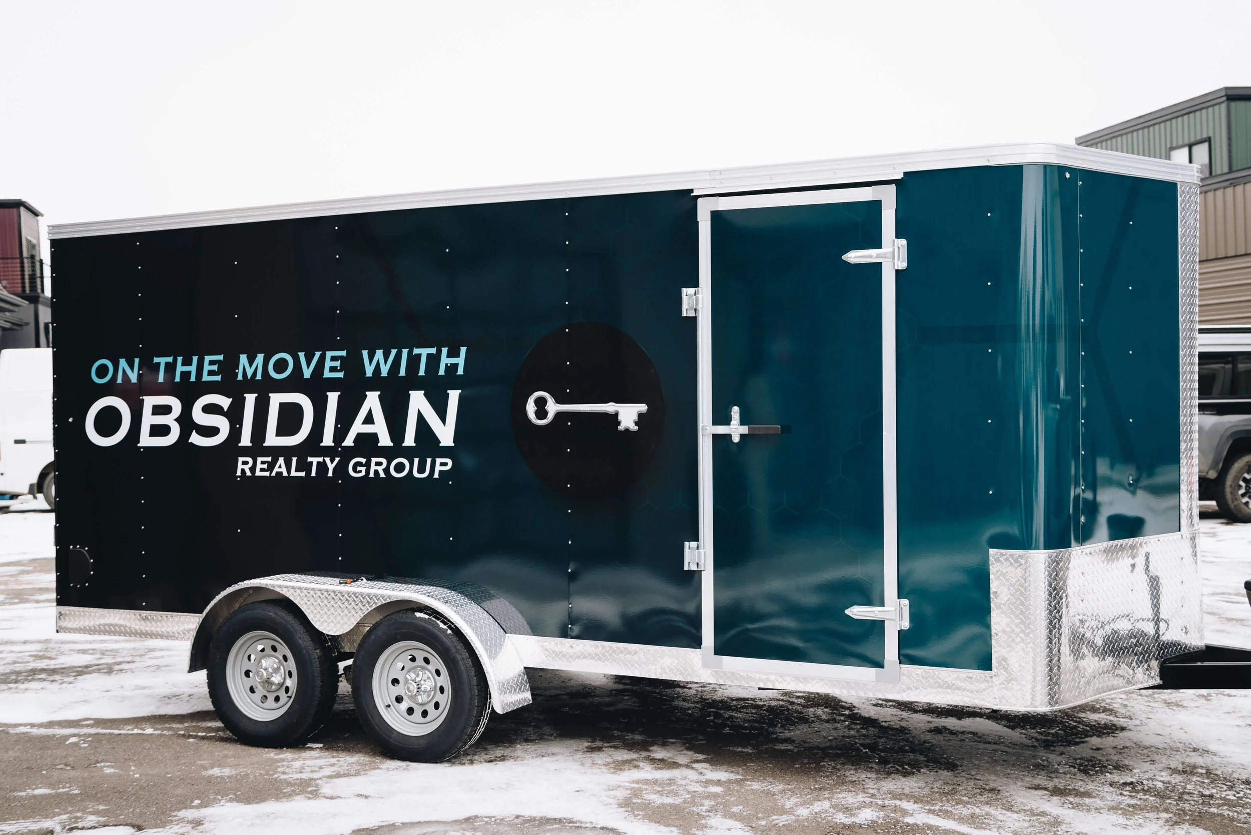 Side view of a dual-axle Obsidian Realty Group trailer in Bozeman, Montana, with sleek dark blue trailer graphics displaying the slogan “On the Move with Obsidian Realty Group” and a key icon.