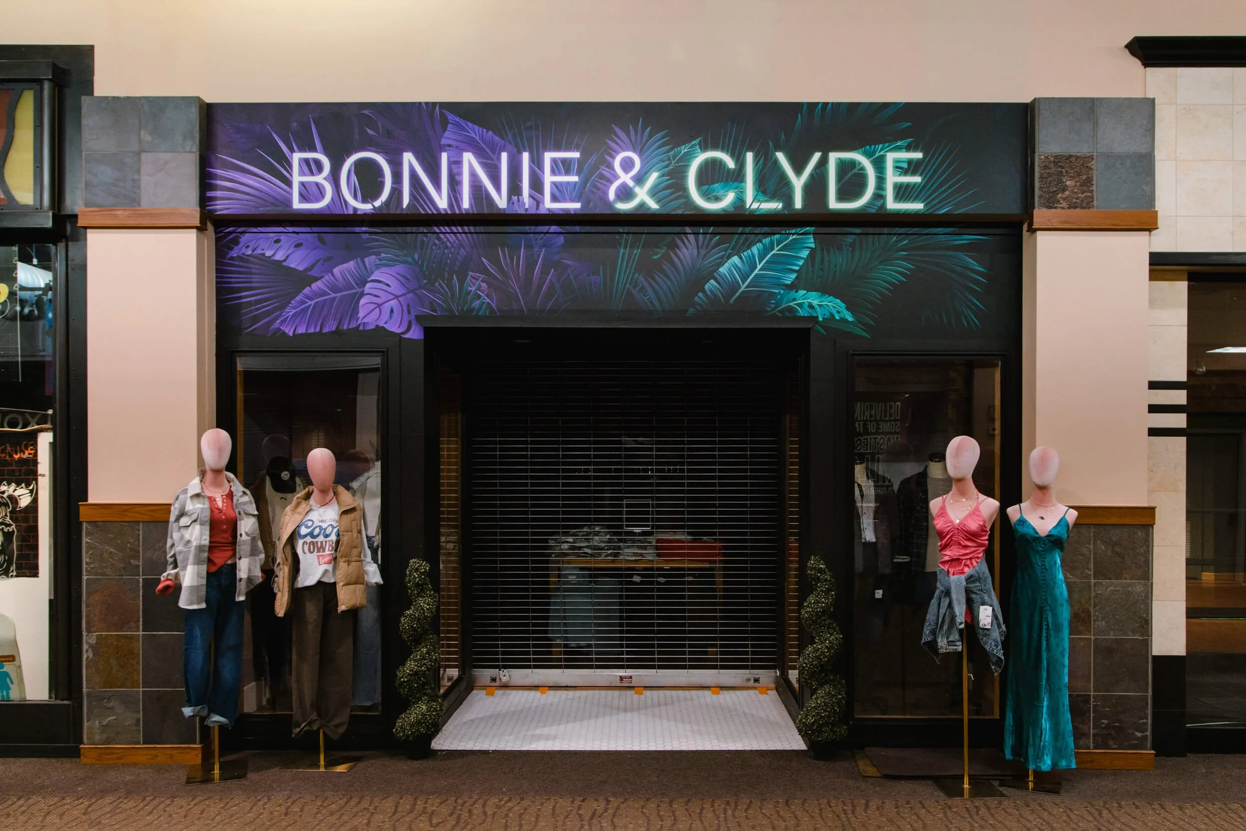 Vibrant retail wall wrap for boutique storefront “Bonnie & Clyde,” featuring neon-style typography and tropical foliage graphics on a dark backdrop, positioned above mannequins at a mall entrance. Business Location: Bozeman, MT
