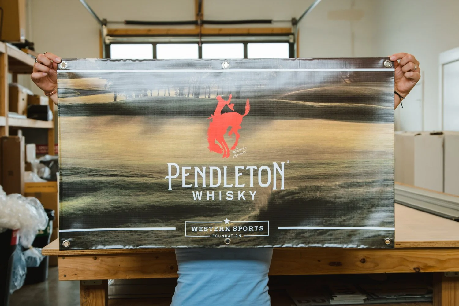Vinyl banner promoting Pendleton Whisky, with scenic background and Western Sports Foundation logo, held in a workshop.
