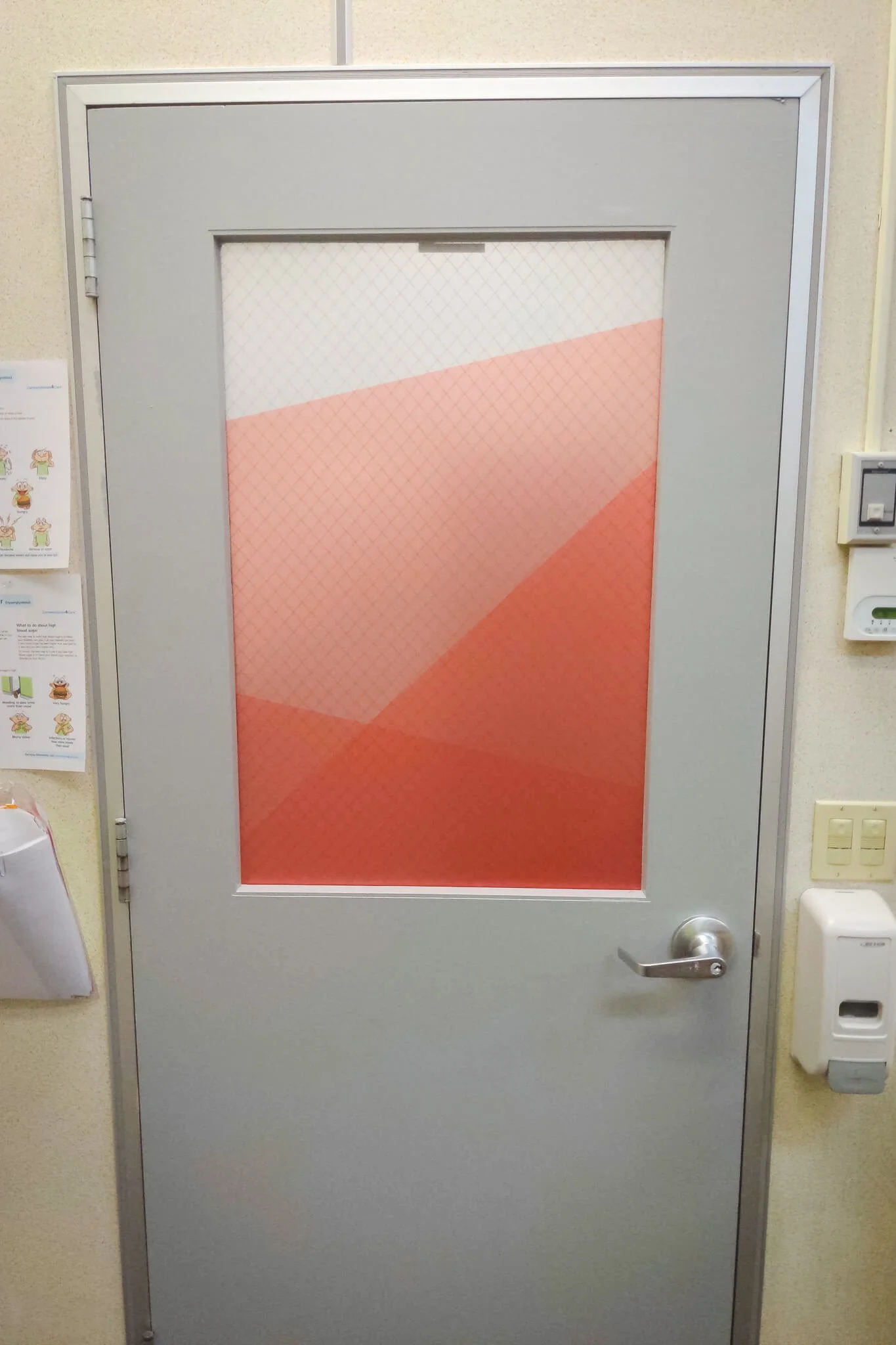 A gray interior door with a rectangular glass panel featuring bold geometric window graphics in red and coral tones, designed as a privacy window film for a school or office setting.