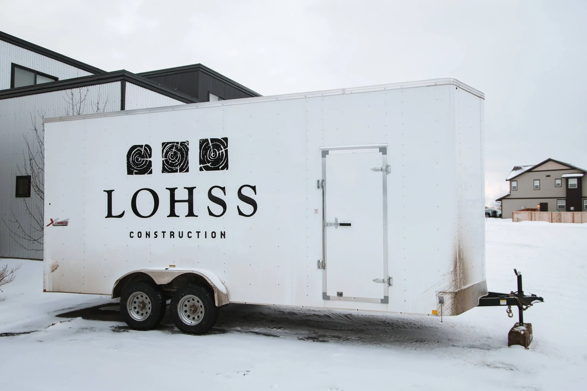 Simple and professional white trailer wrap for LOHSS Construction in Bozeman, Montana, with bold black logo and stylized tree ring graphics, parked on a snowy site.