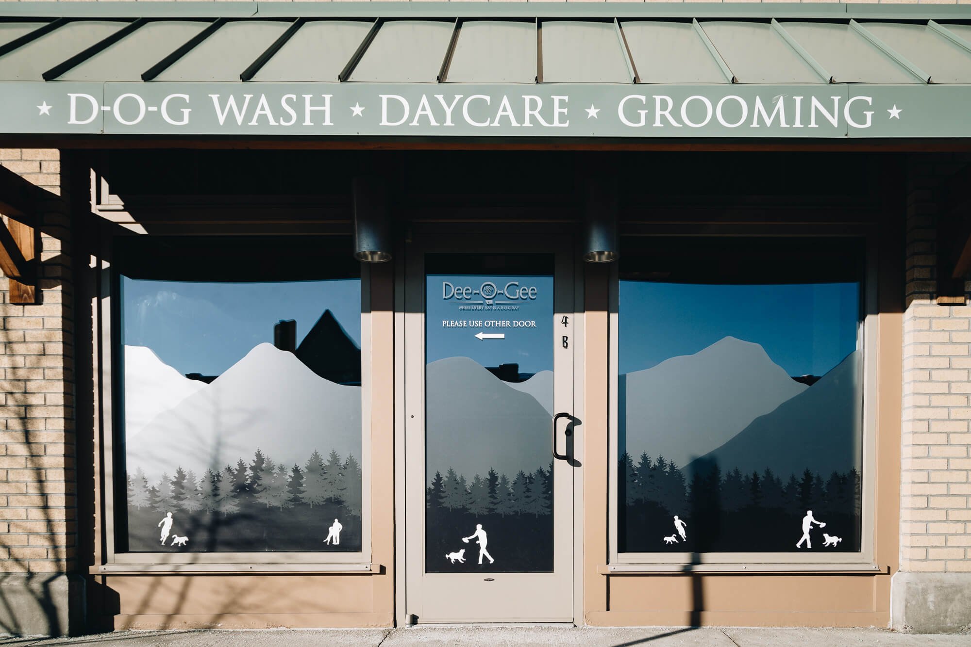 Full storefront window graphics at Dee-O-Gee showcase a mountain and forest silhouette with people walking dogs, under signage for wash, daycare, and grooming.