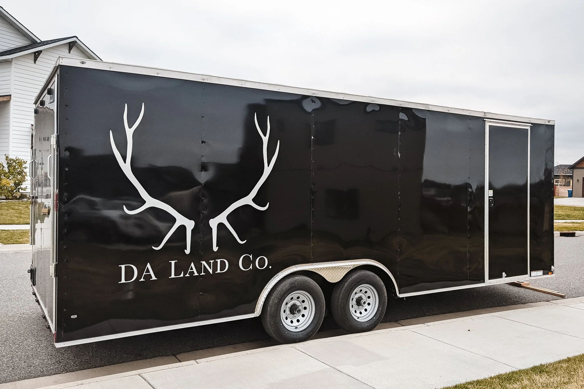 DA Land Co. trailer graphics in Bozeman, Montana, featuring a black wrap with large white antler artwork and serif business name text on a tandem axle trailer parked on a residential street.