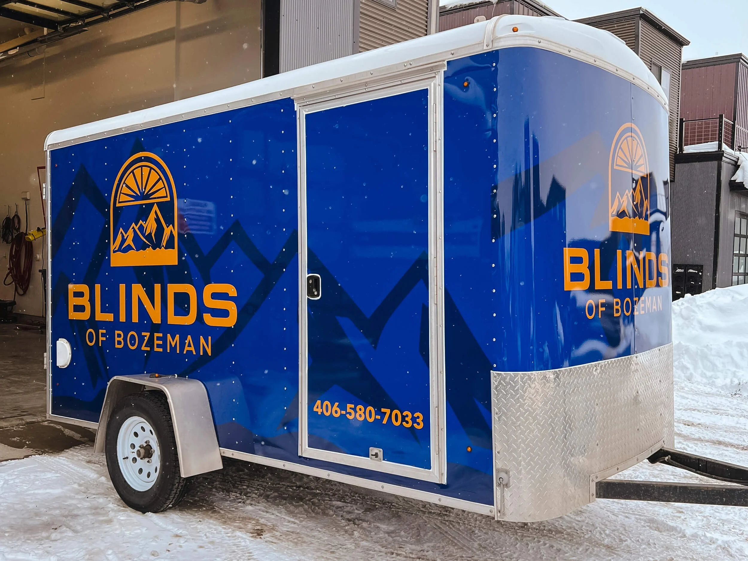 Bright blue trailer wrap with a watermarked mountain pattern overlaid with bright gold Blinds of Bozeman logos and contact information