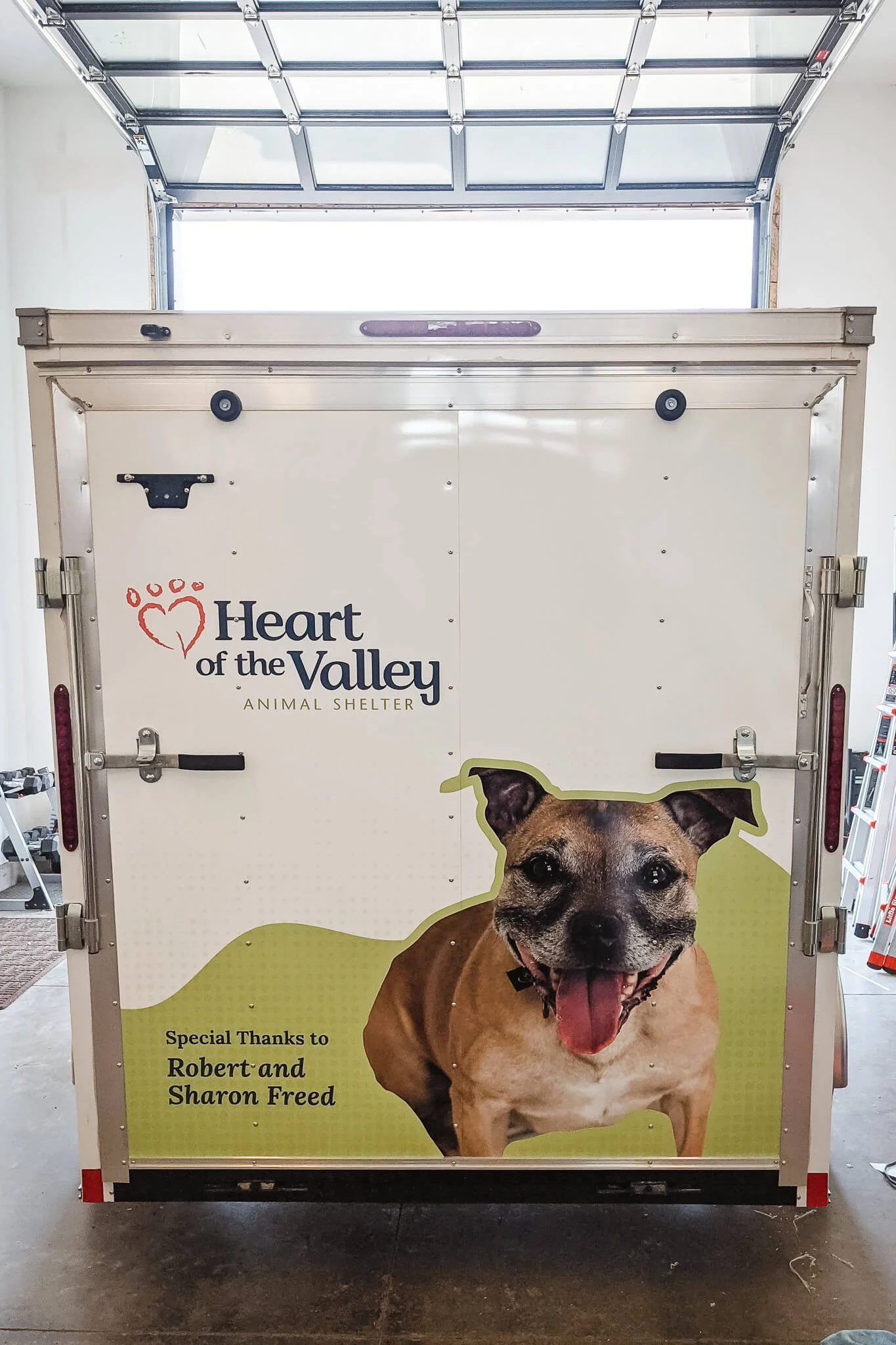 Rear view of a white enclosed trailer featuring Heart of the Valley Animal Shelter trailer graphics in Bozeman, Montana, with a large photo of a smiling dog and donor recognition text, ideal for mobile branding and nonprofit visibility.