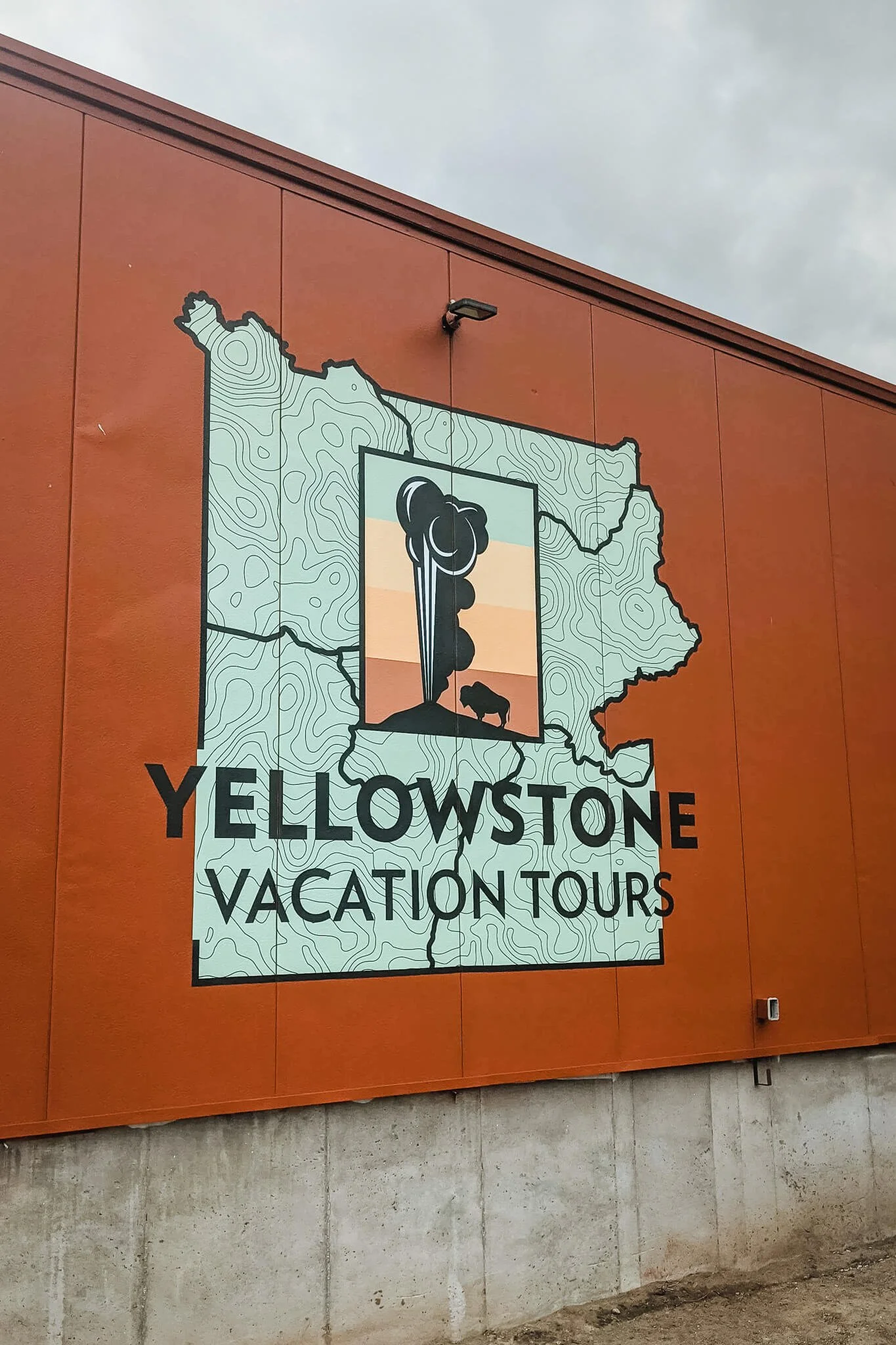 Exterior orange building wall featuring a large custom wall wrap with the Yellowstone Vacation Tours logo, map design, and illustration of a geyser and bison in West Yellowstone, MT.
