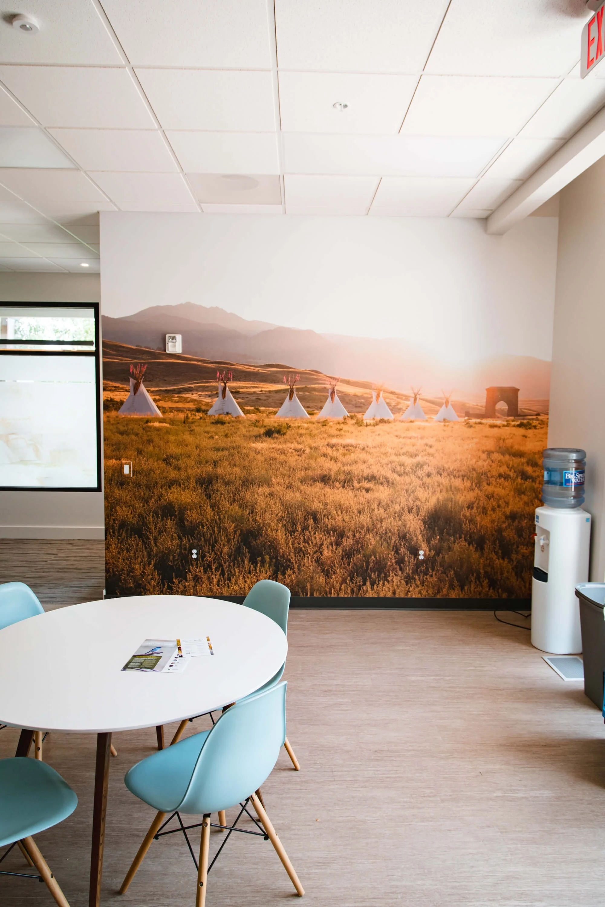 Scenic wall wrap showcasing a row of teepees in a glowing sunset over golden fields, enhancing a casual seating area with cultural and natural beauty. Installed at Yellowstone Forever in Bozeman, MT.