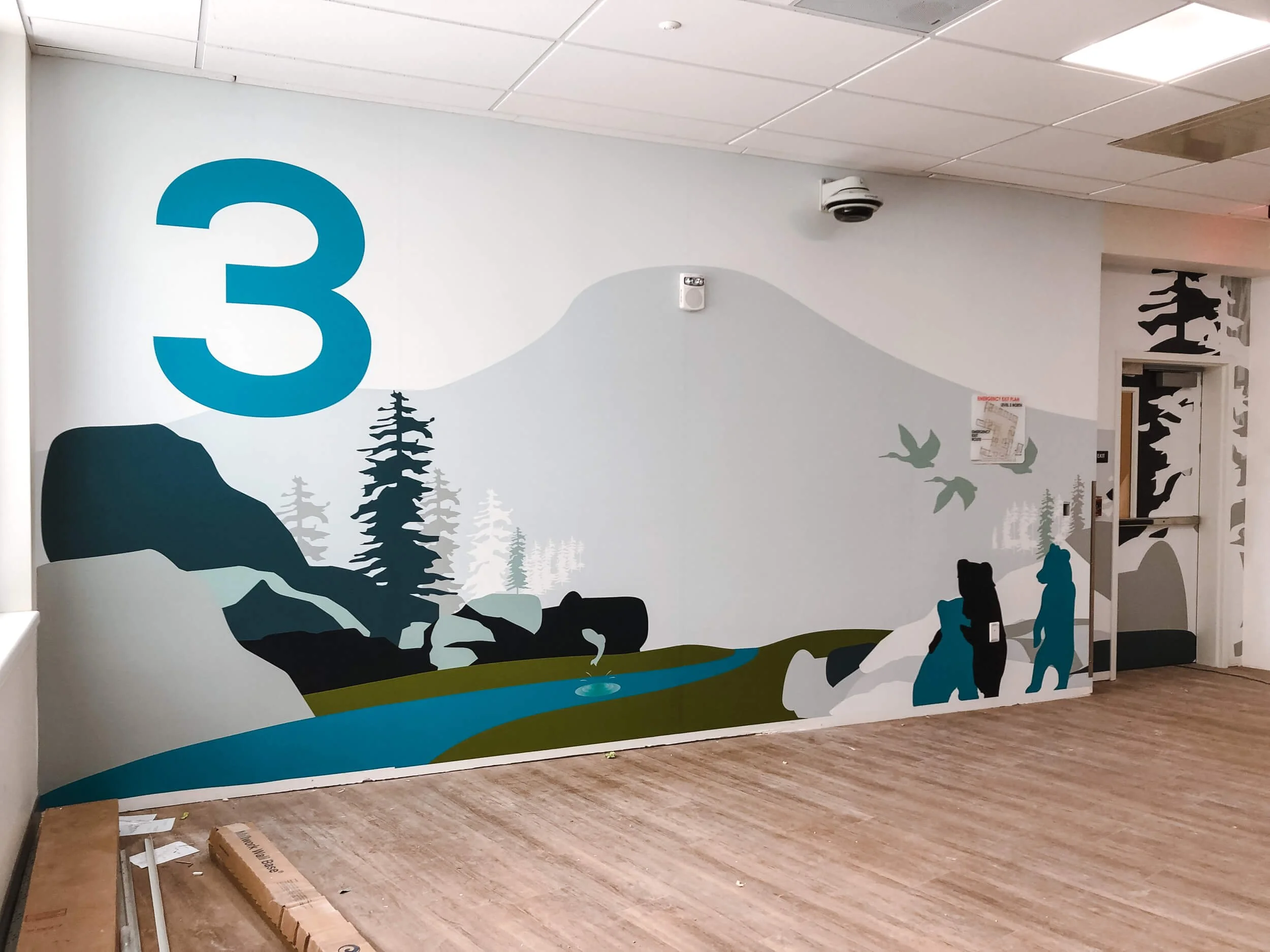 Illustrative forest and wildlife wall wrap with a large blue number 3, featuring silhouetted bears, pine trees, and a stream, used as a wayfinding or floor identification marker. Installed at Shodair Children’s Hospital in Helena, MT.