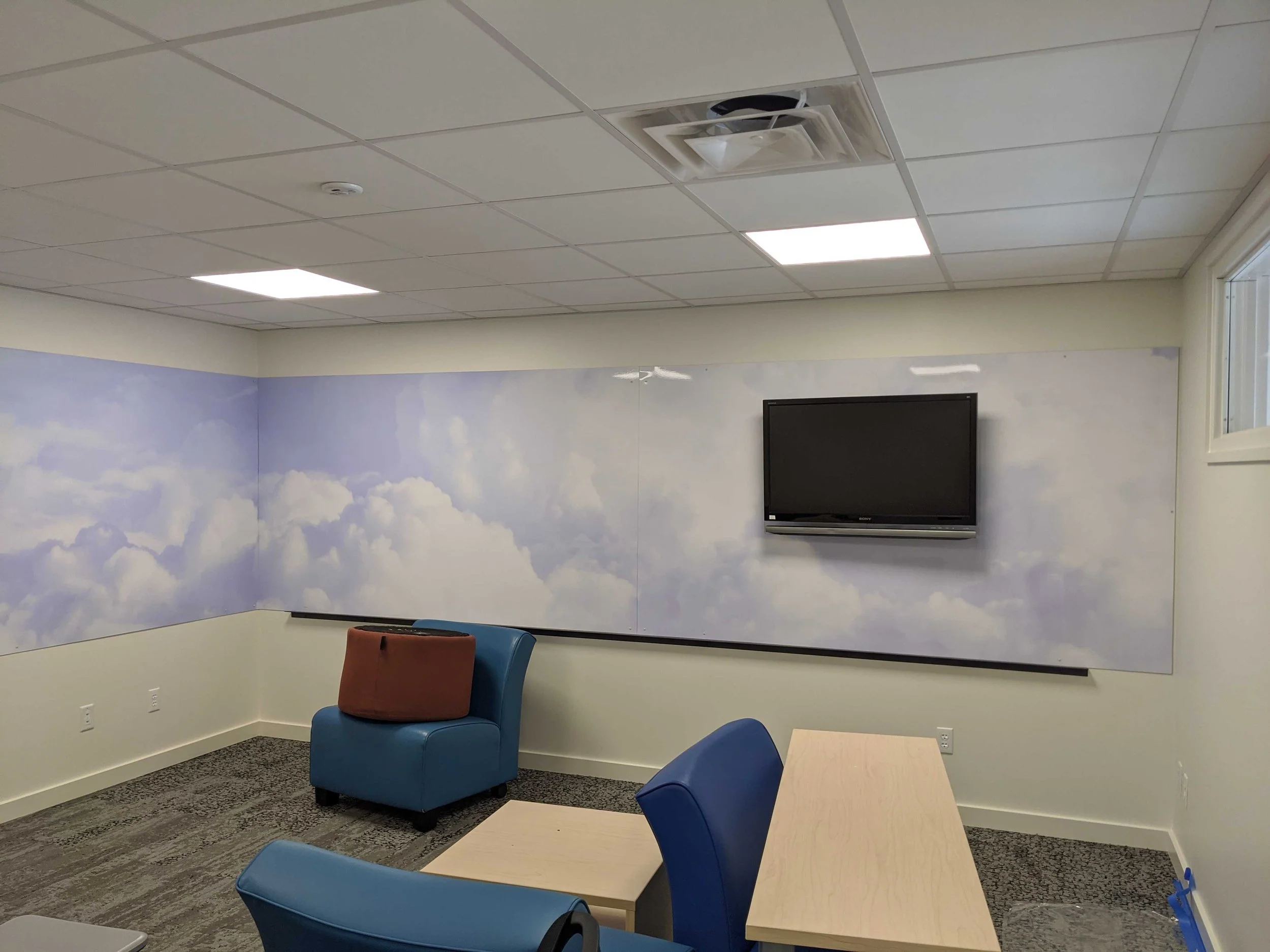 Cloud-patterned dry erase wall graphics wrapping around a corner above blue seating.