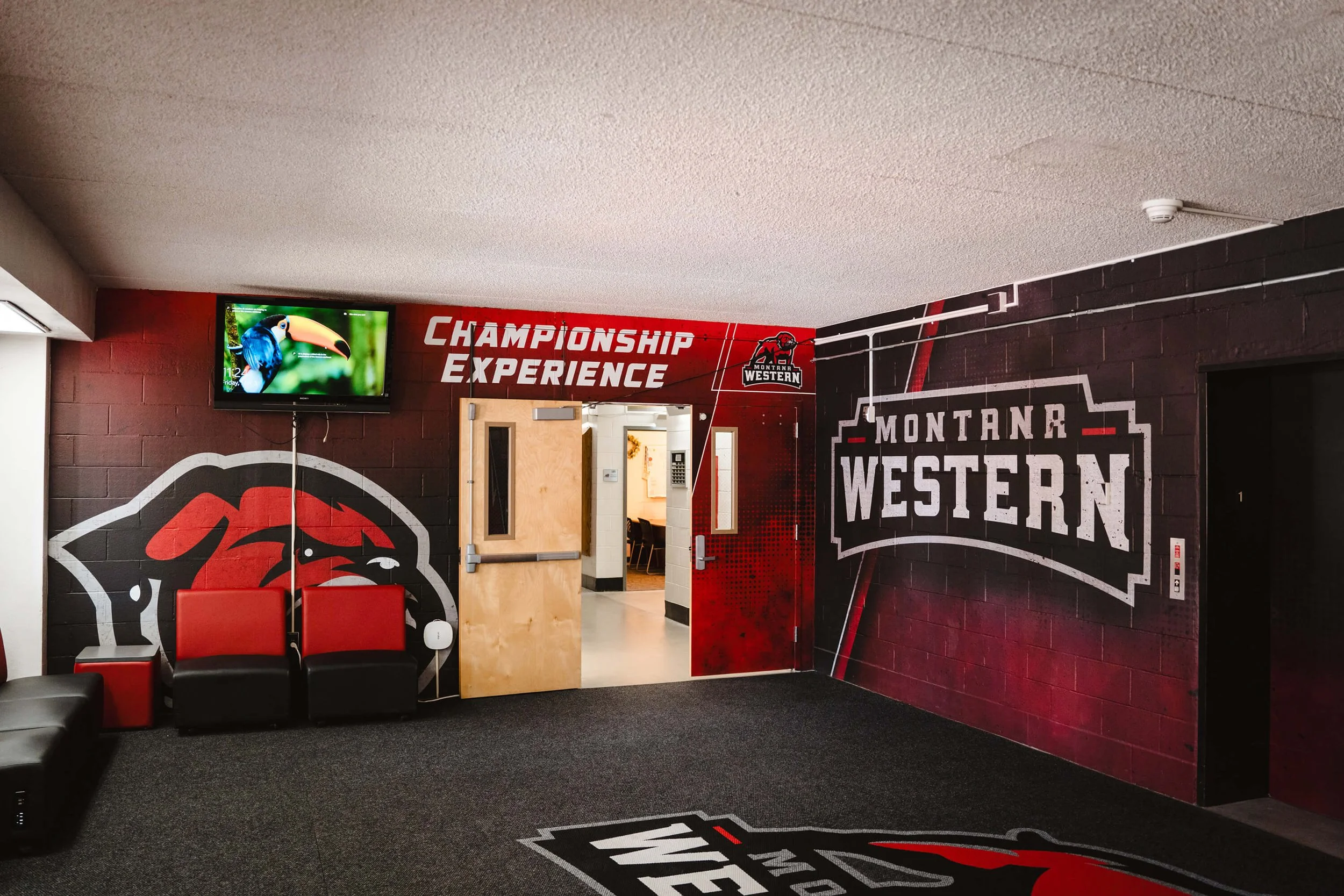 Entrance area with red and black Montana Western wall wraps showcasing the bulldog mascot, “Championship Experience” text, and school logo near seating and open double doors in Dillon, MT.