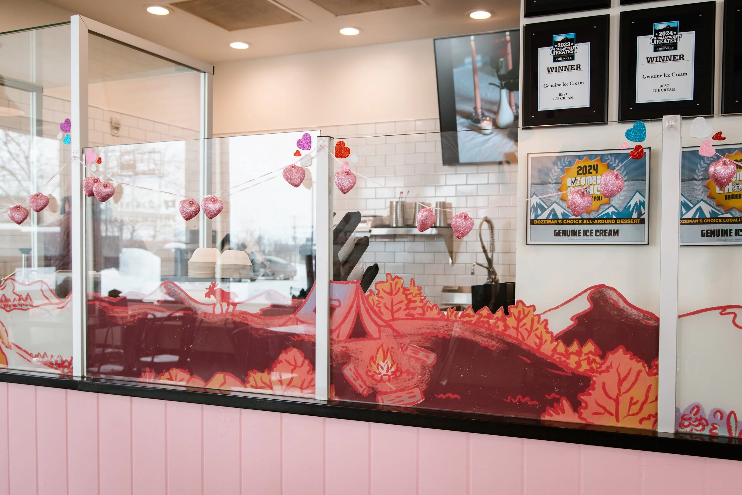 Decorated glass panel inside Genuine Ice Cream in Bozeman, MT, with red-toned window graphics of a mountain campsite scene, complete with trees and a tent.