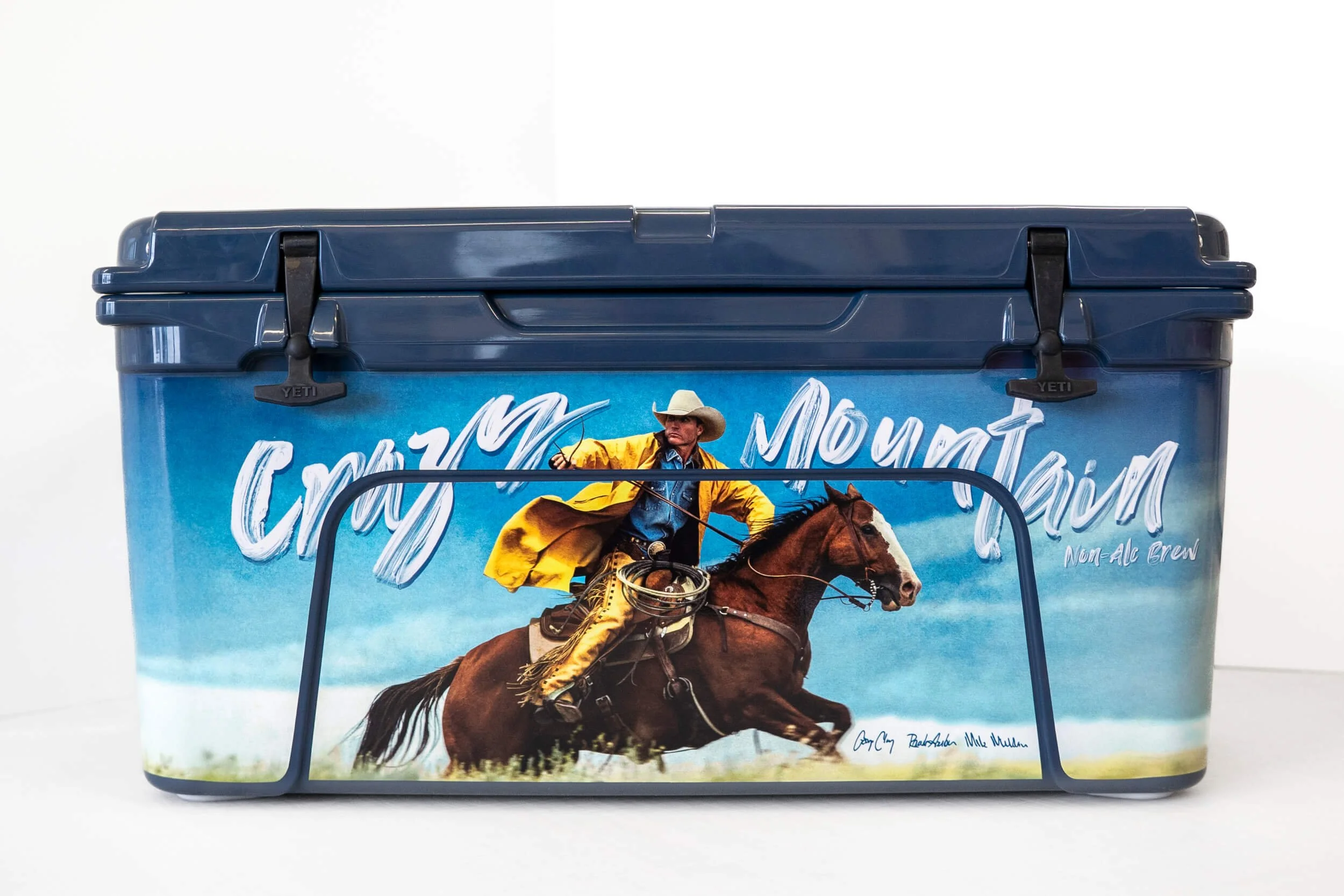 Crazy Mountain text arcing above a cowboy wearing a yellow rain jacket riding a horse on a custom vinyl wrapped YETI cooler