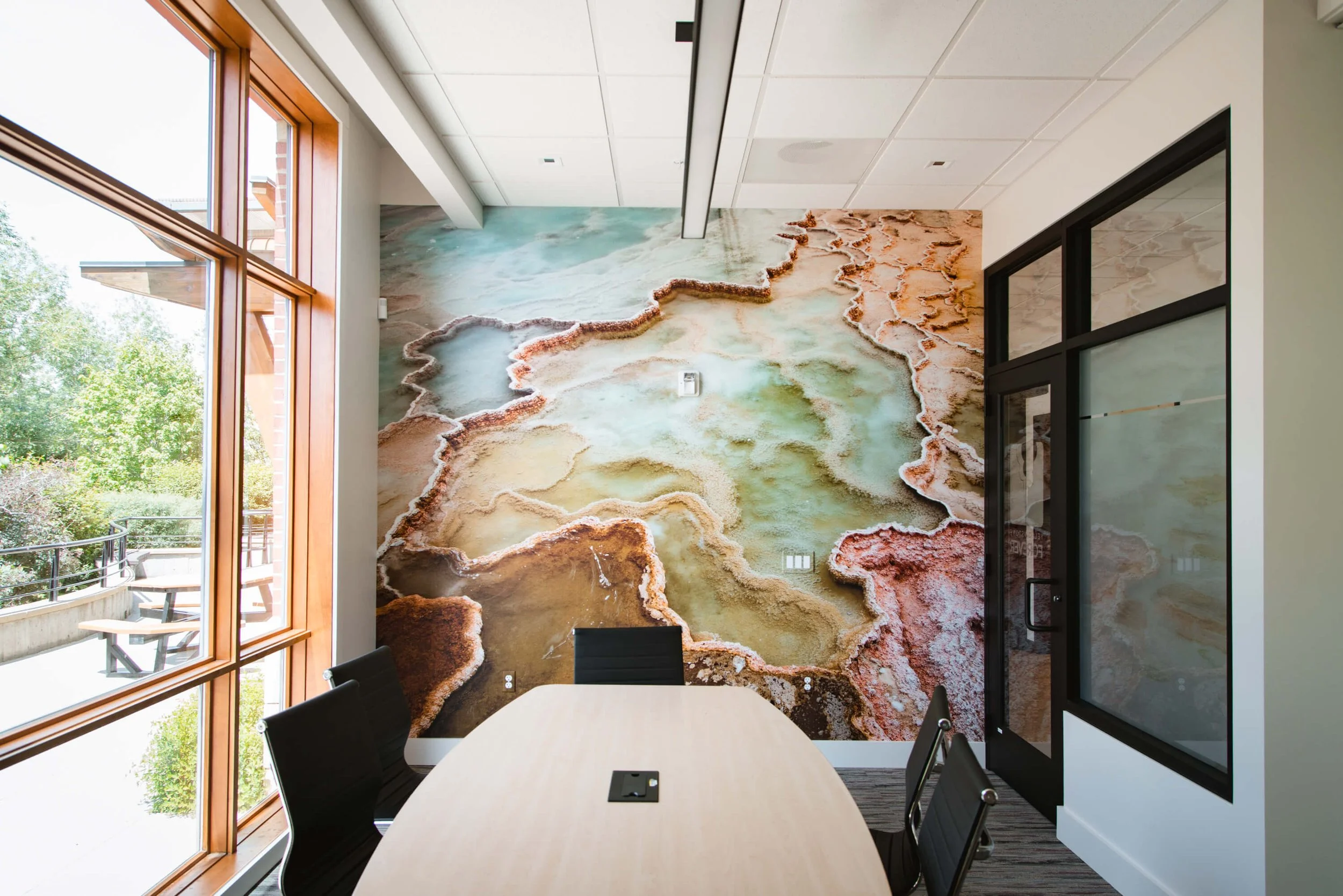 Artistic wall wrap depicting an aerial view of a mineral-rich landscape with vibrant blues and earthy tones, creating a serene, nature-inspired feature wall in a modern conference room. Installed at Yellowstone Forever in Bozeman, MT.