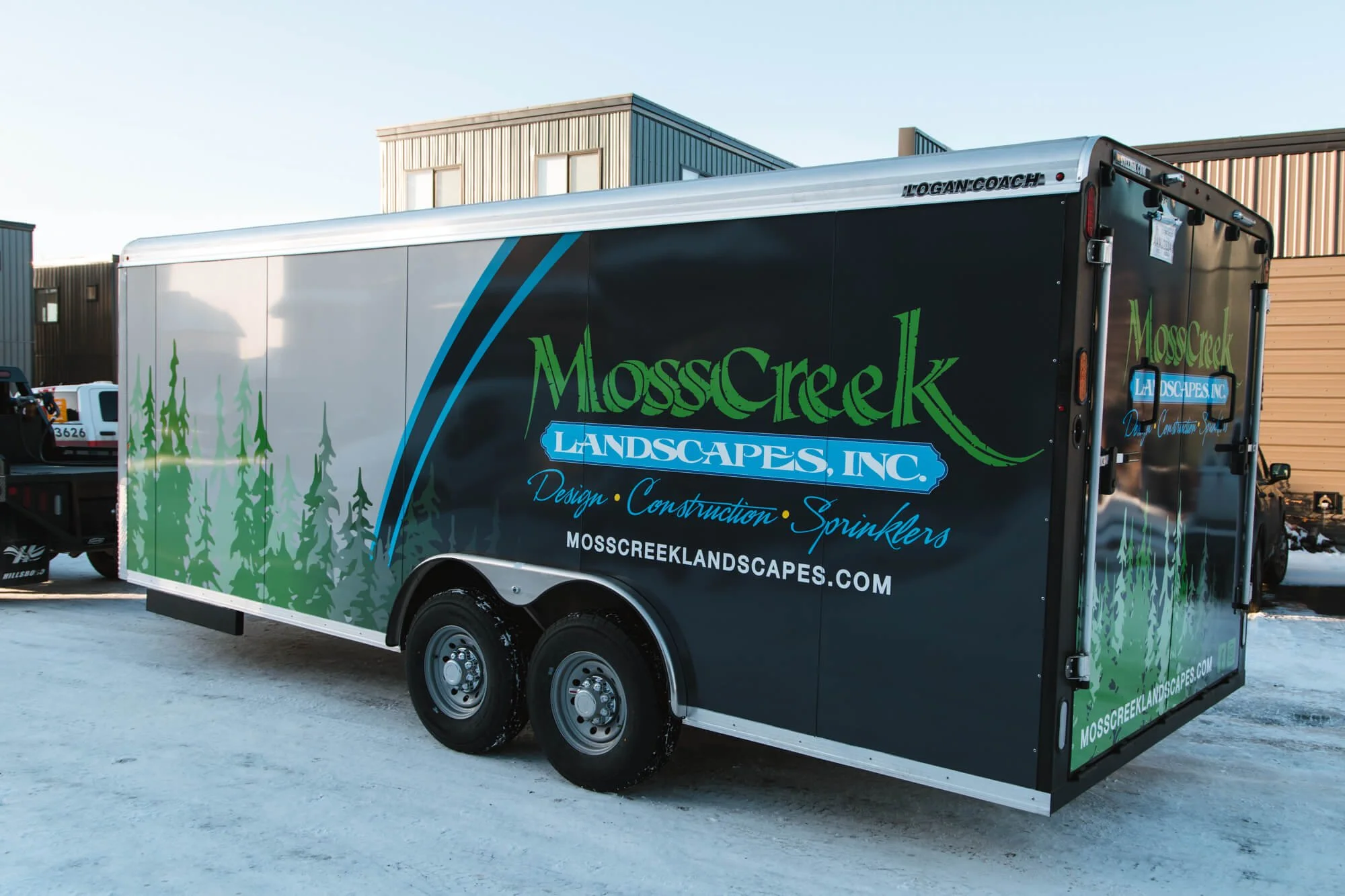 MossCreek Landscapes trailer graphics in Bozeman, Montana, featuring green evergreen trees and a bold logo on a deep blue wrap, highlighting design, construction, and sprinkler services.