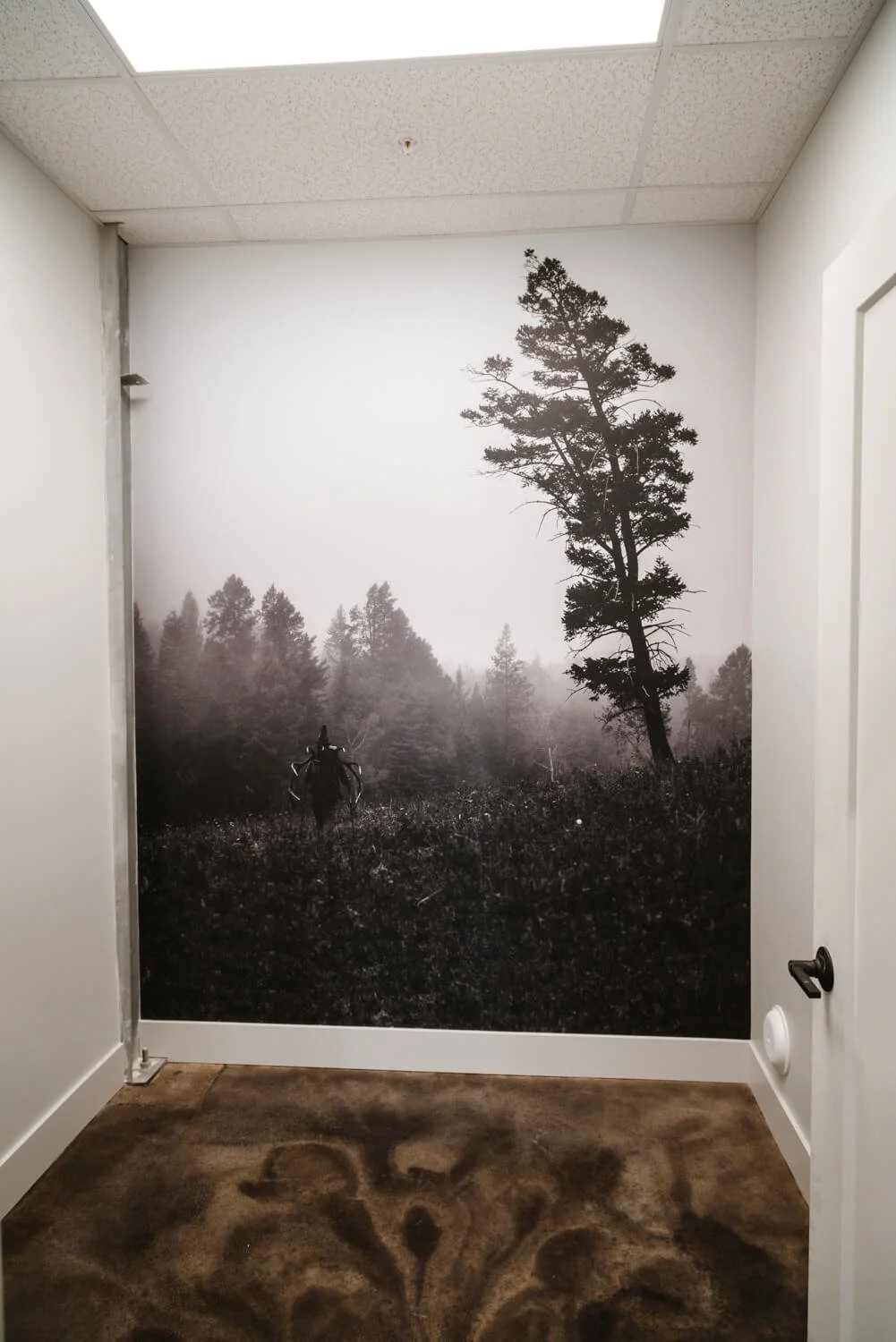Black and white wall wrap featuring a dramatic forest scene with a silhouetted figure carrying antlers, adding a rugged and rustic feel to an office hallway. Installed at Stone Glacier in Bozeman, MT.