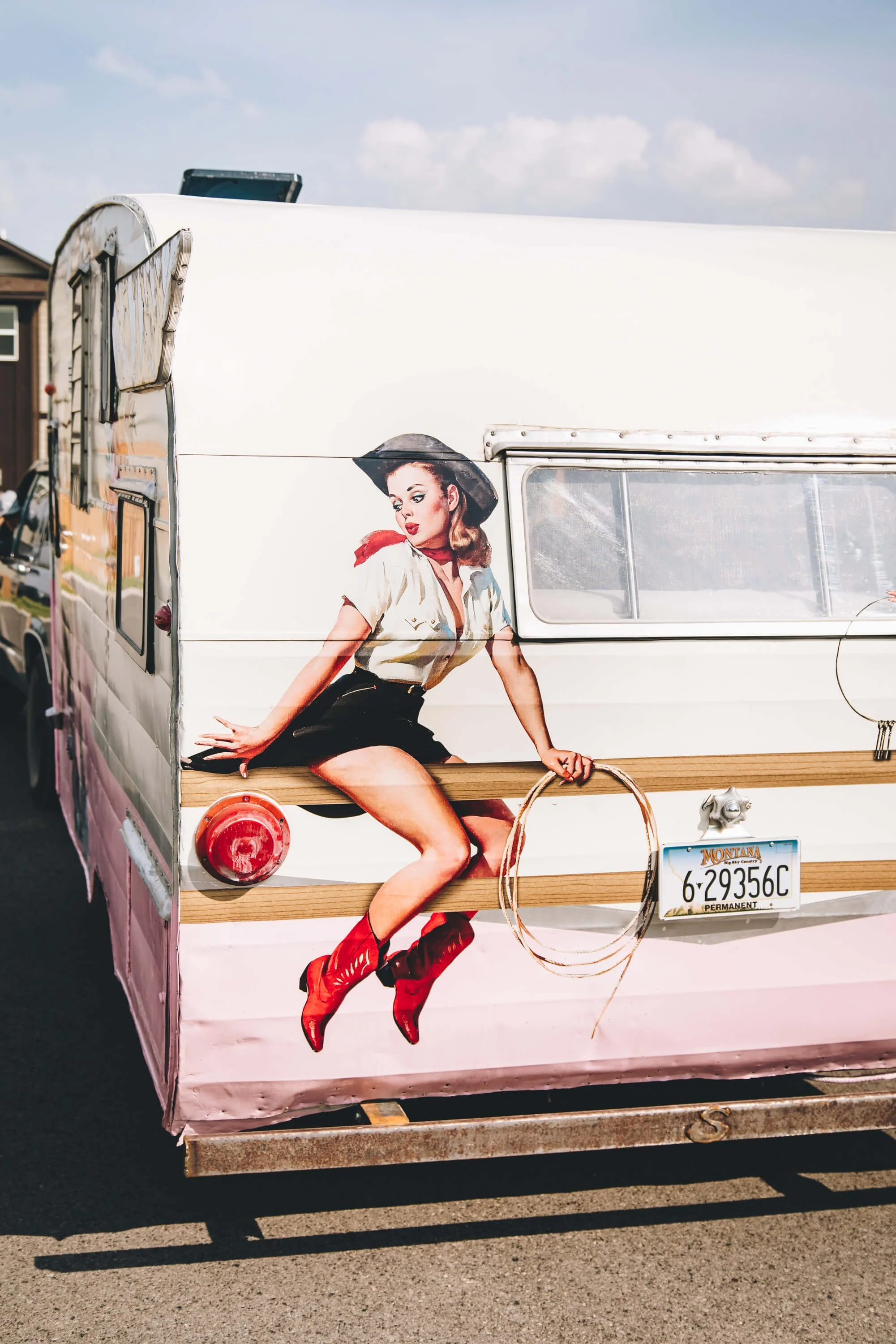 Vintage camper trailer wrap featuring a retro-style cowgirl in red boots and a lasso, printed on the rear panel with a painted wood rail detail.