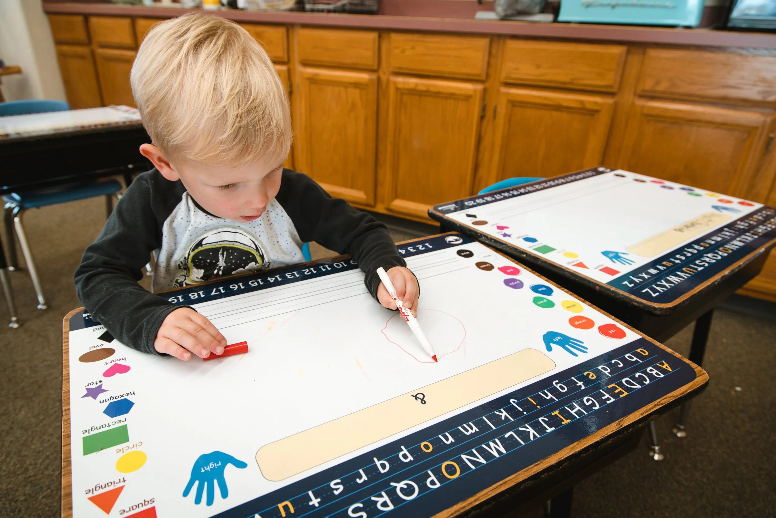 Young student drawing on a custom dry erase desk wrap bordered by alphabet letters and colorful learning graphics.
