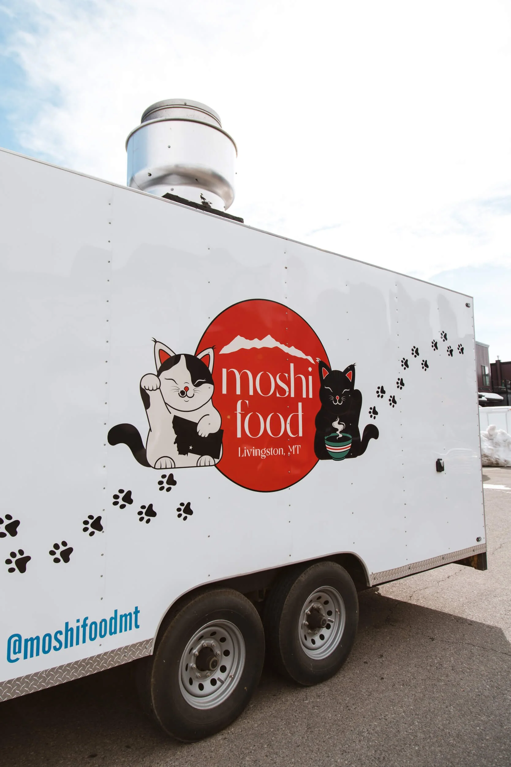 White trailer with minimalist Moshi Food graphics, including waving cat mascots, paw print trails, and logo for the Livingston, MT-based food truck. 