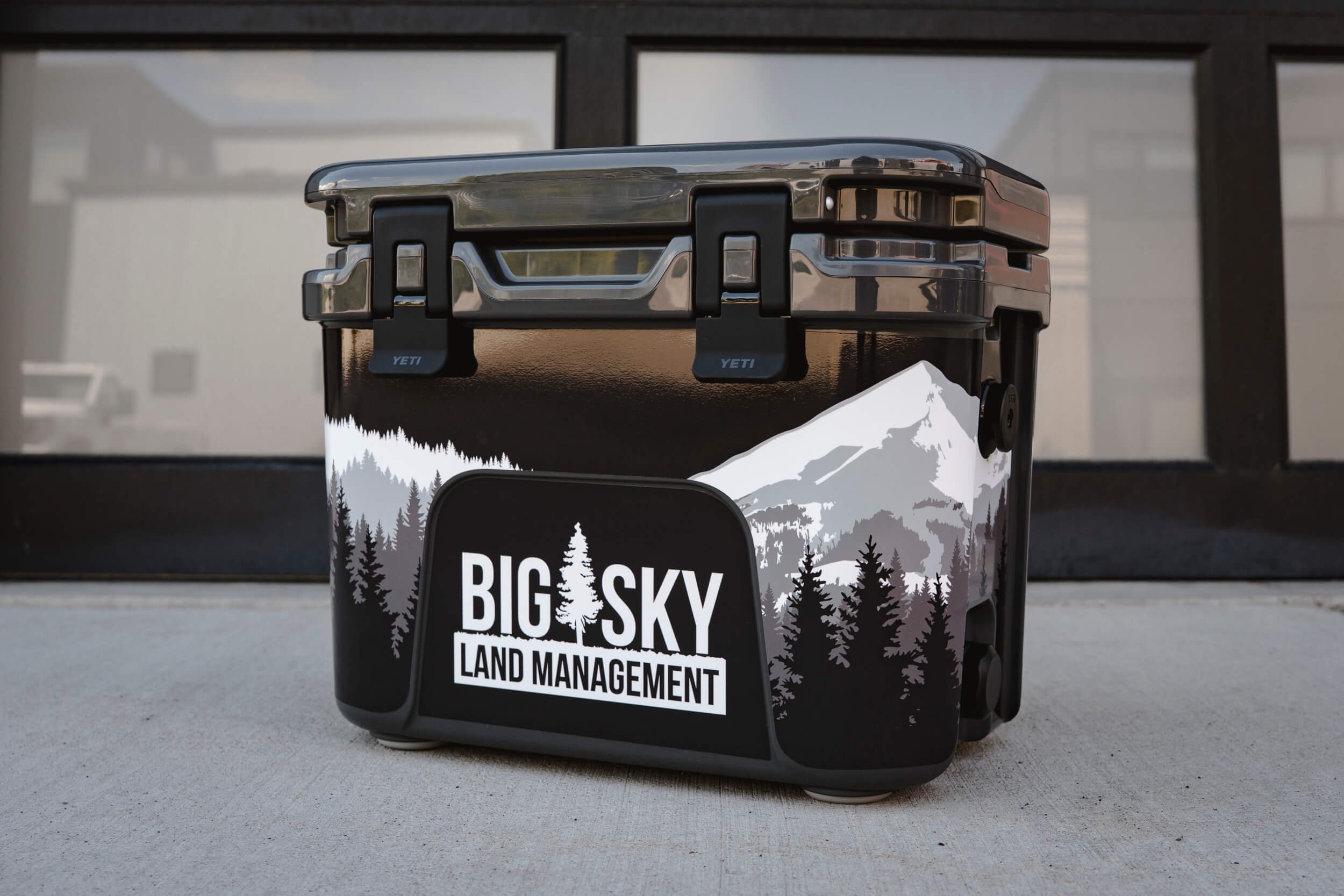 Small charcoal YETI cooler with a Big Sky Land Management logo on the center front with a white and grey illustration of Lone Peak in the background. Local branding