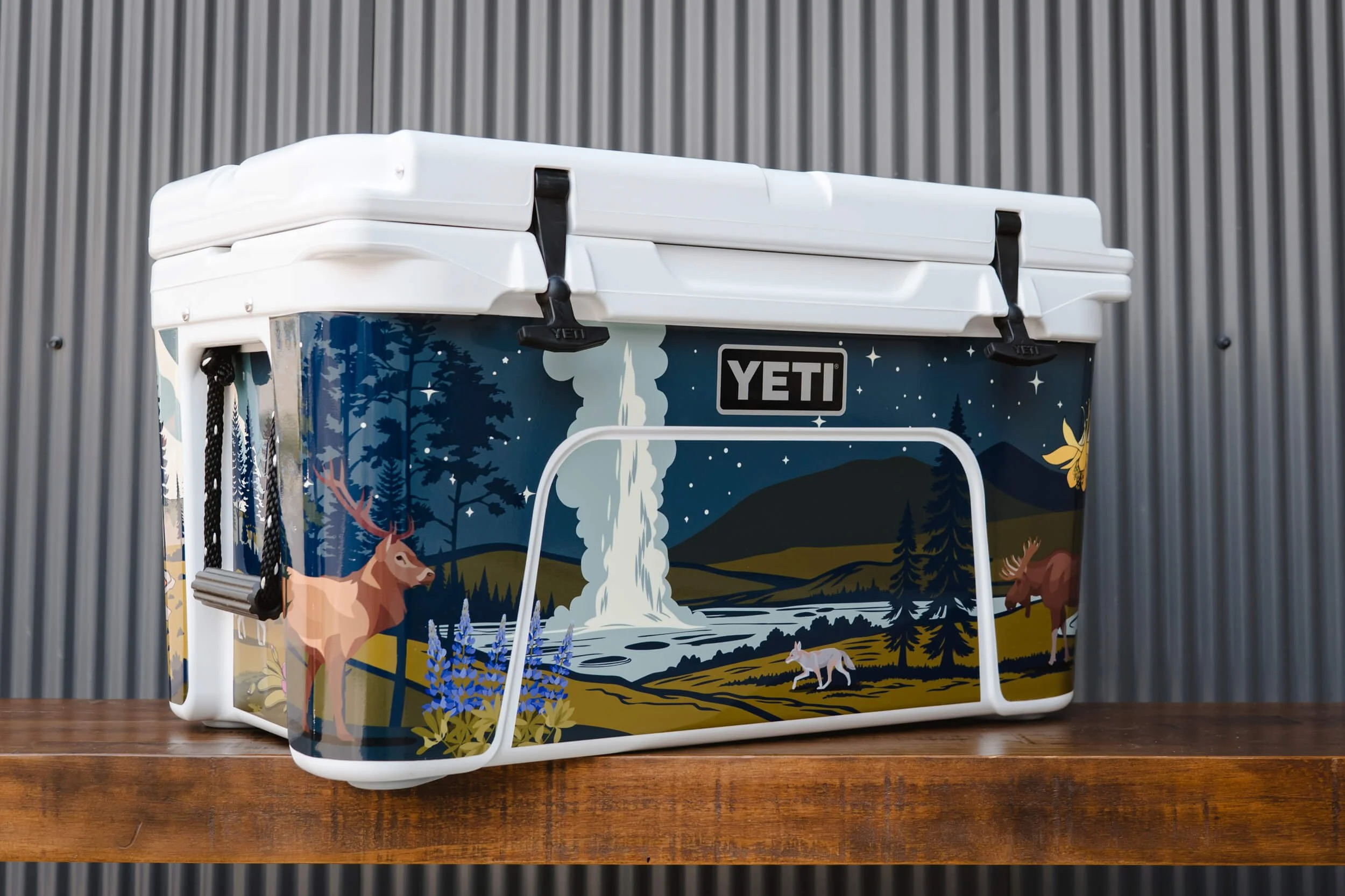 Angled photo of a YETI cooler with an elk, coyote and moose with Old Faithful in the background on a vinyl wrap with a Yellowstone National Park theme