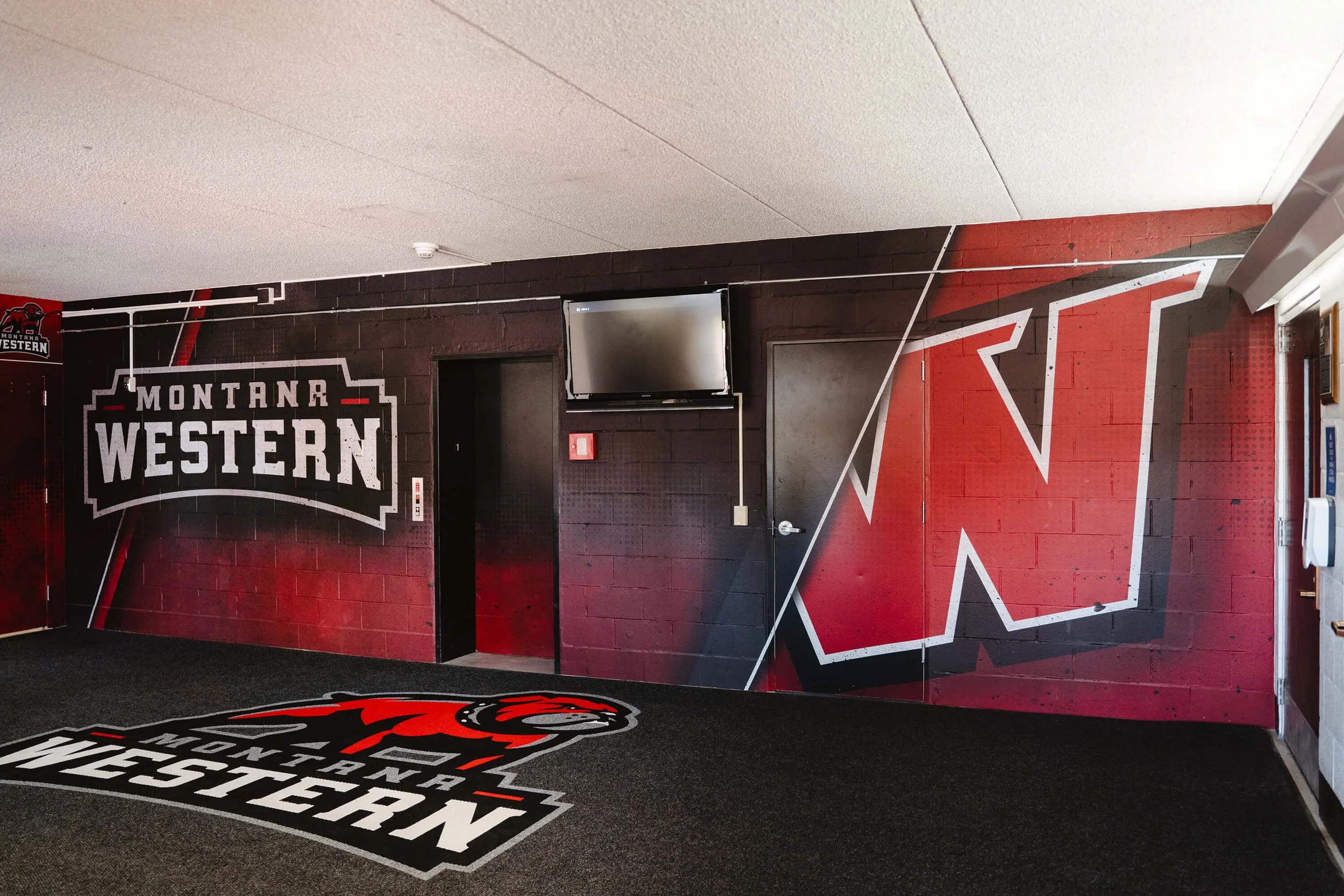 Interior athletic facility featuring a bold red and black Montana Western wall wrap with a large “W” logo and school branding on cinderblock walls, adding school pride and professional design in Dillon, MT.