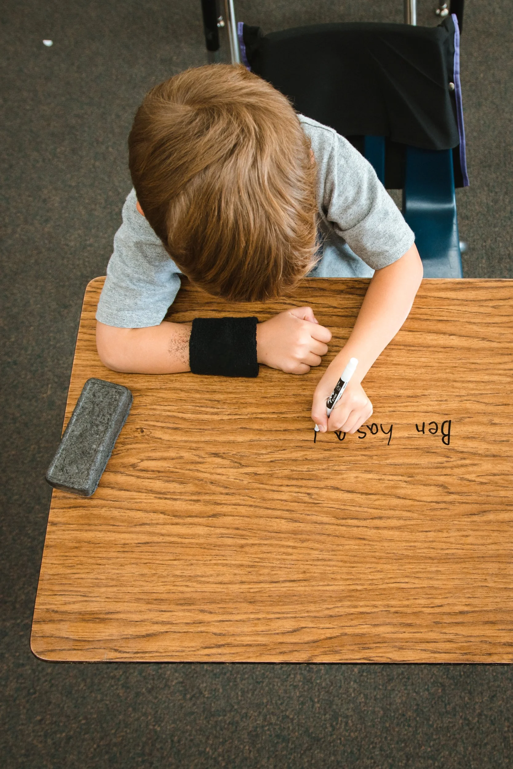 Student writing a sentence on a woodgrain dry erase desktop with an eraser beside it.