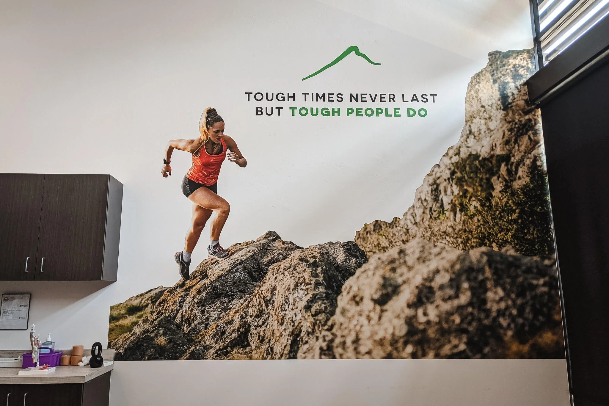 Vinyl wall spot graphic of a woman in athletic gear climbing rocky terrain with motivational quote 'Tough times never last but tough people do' on wall.