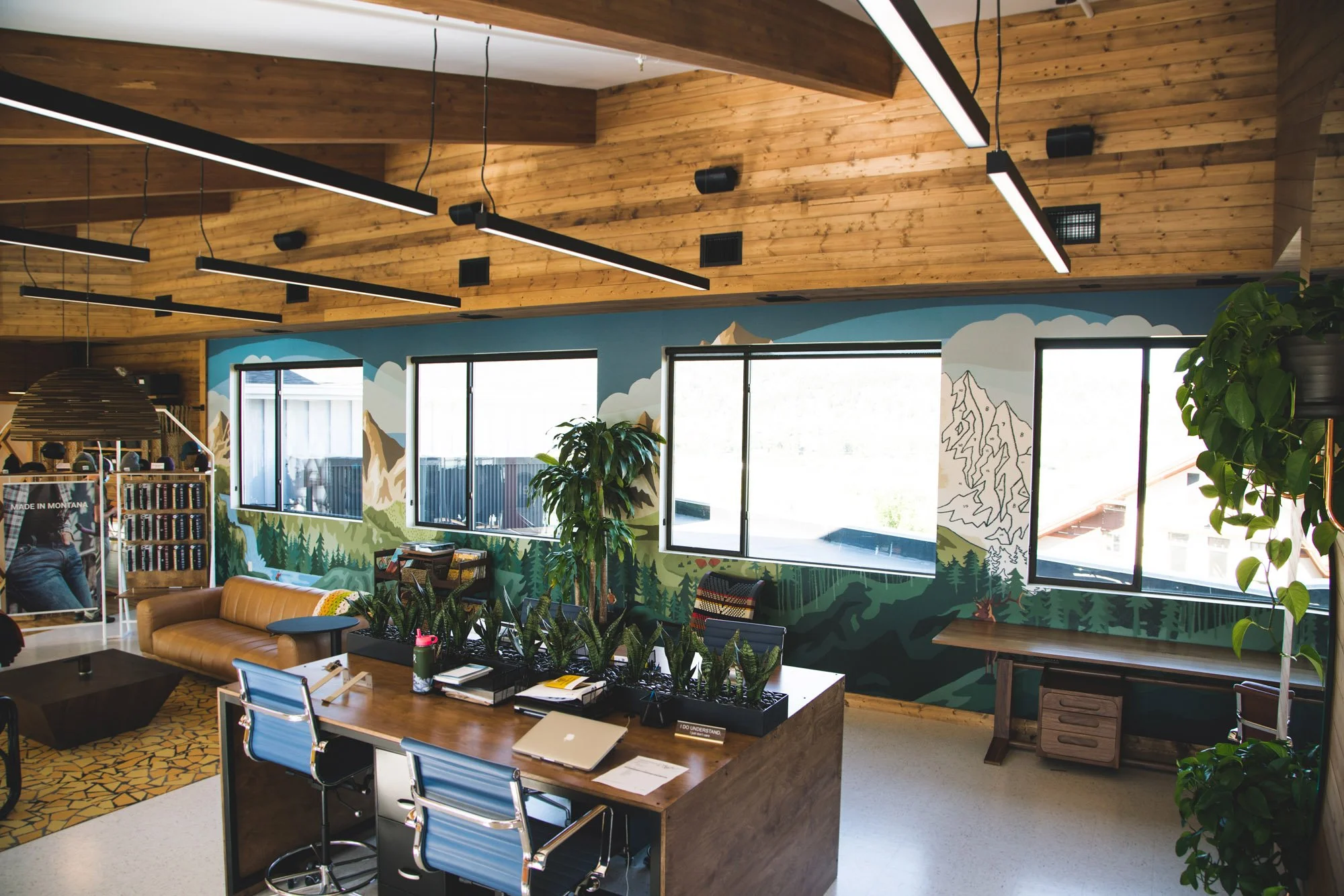 Panoramic wall wrap of layered mountain ranges and pine forests stretching across an open-concept office wall with large windows at formerly Jelt Belt in Bozeman, MT.
