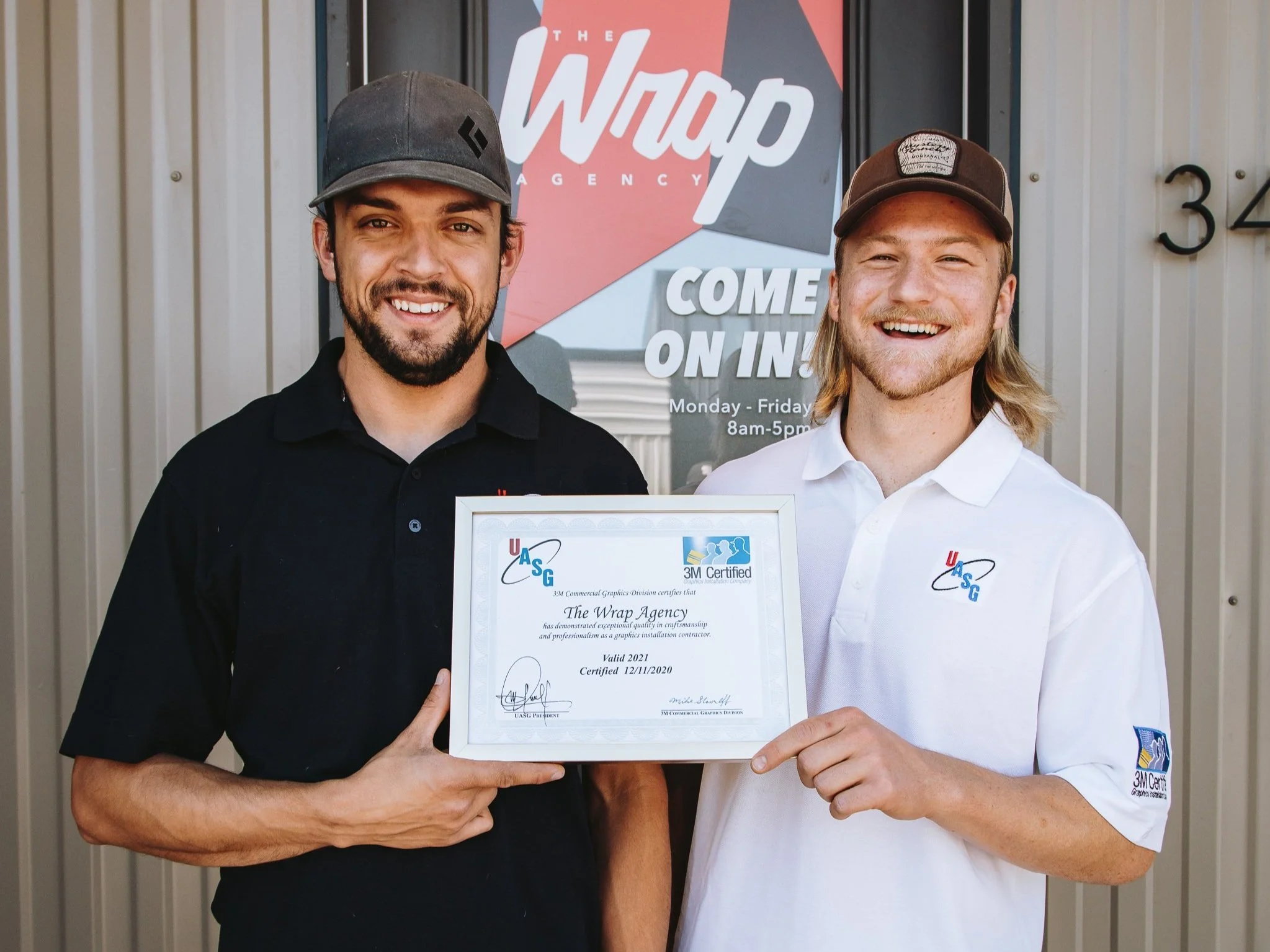 Two installers standing outside holding a certification plaque with "3M Certified" and "UASG" logos, smiling at the camera. A sign behind them reads "The Wrap Agency."