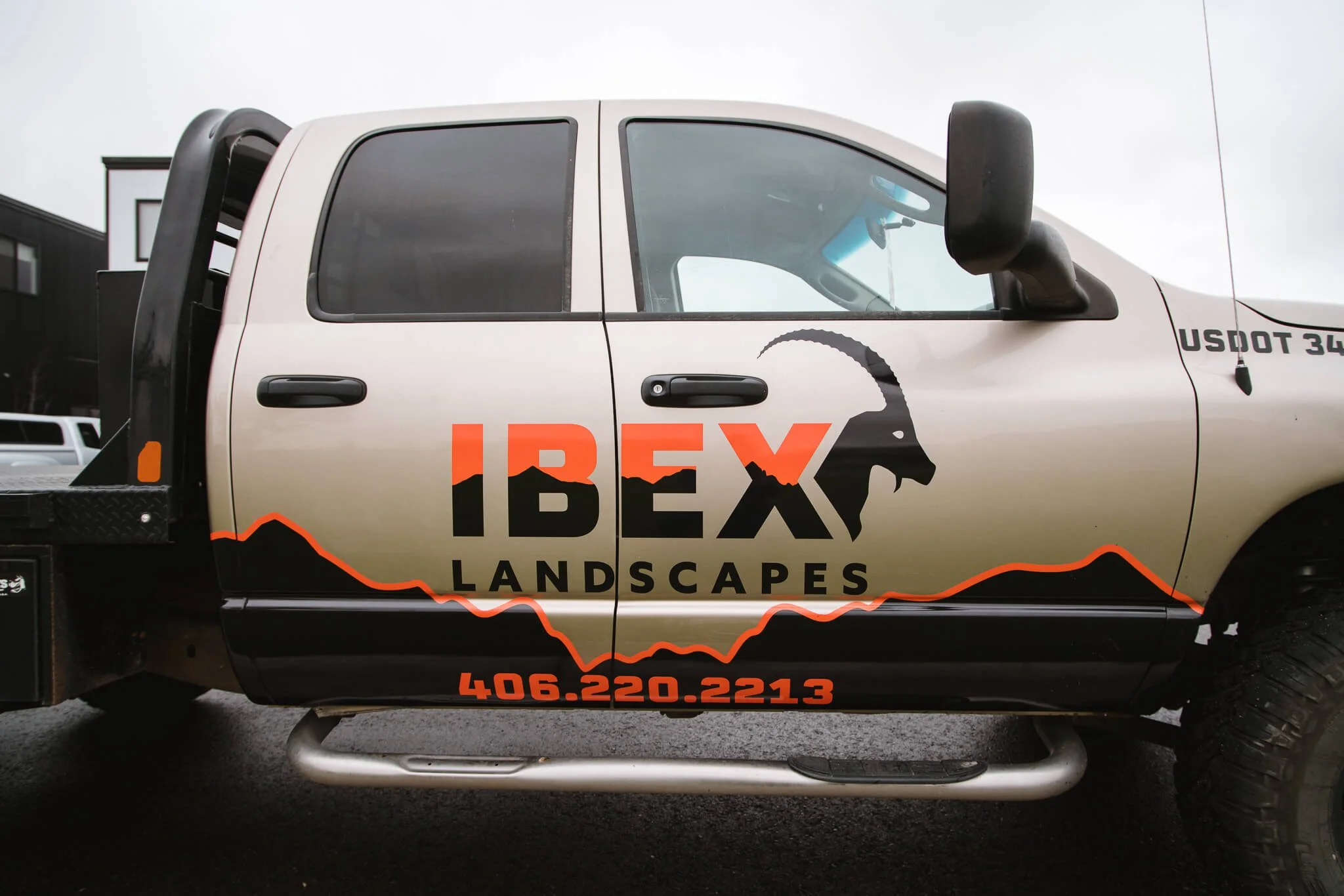 Tan and black pickup truck in Bozeman, MT, with a bold IBEX Landscapes vehicle wrap featuring a mountain range graphic and mountain goat logo in orange and black vinyl.