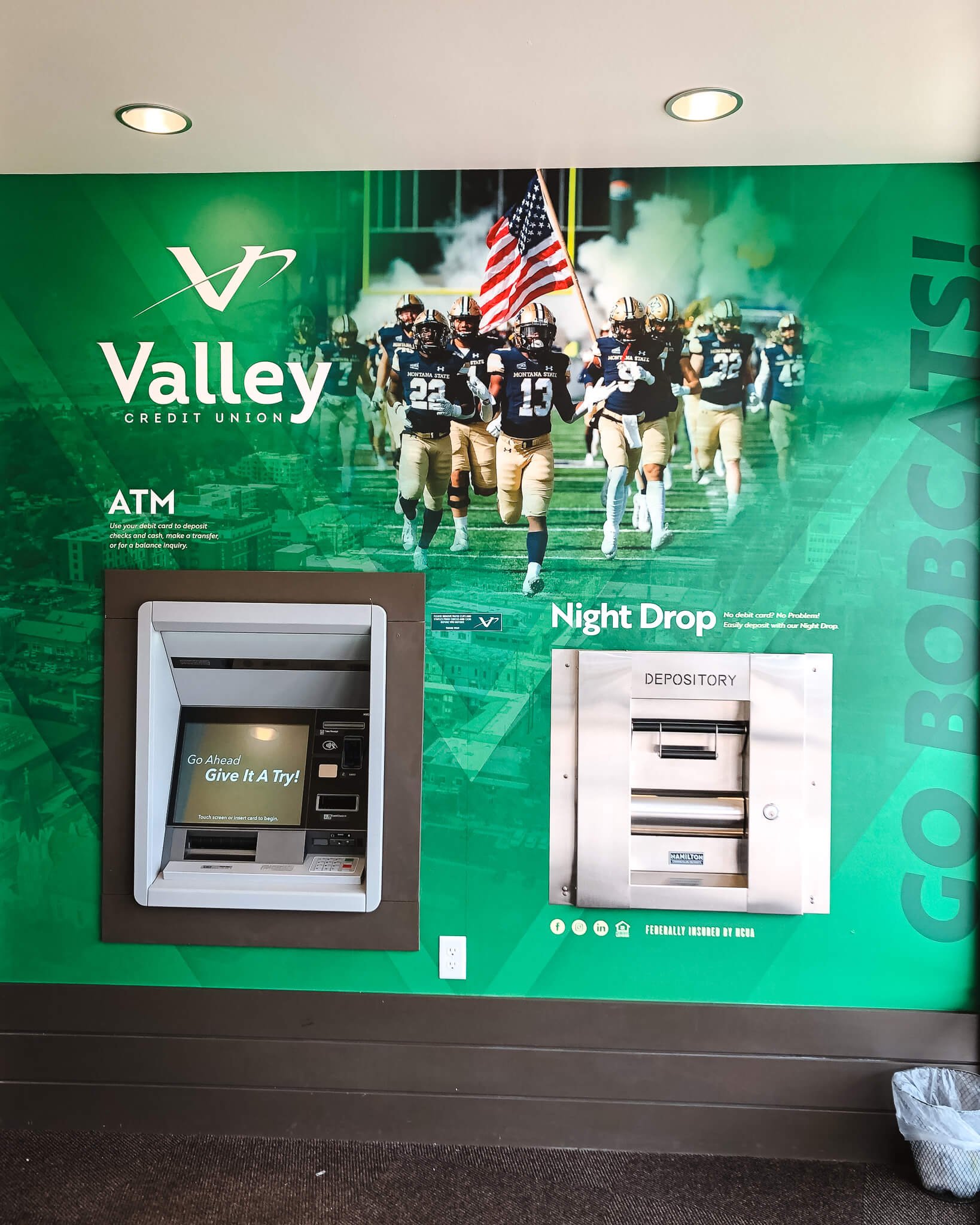 Eye-catching wall wrap surrounding ATM and night drop stations, featuring a photo of college football players running with a flag and branding for Valley Credit Union in Bozeman, MT.