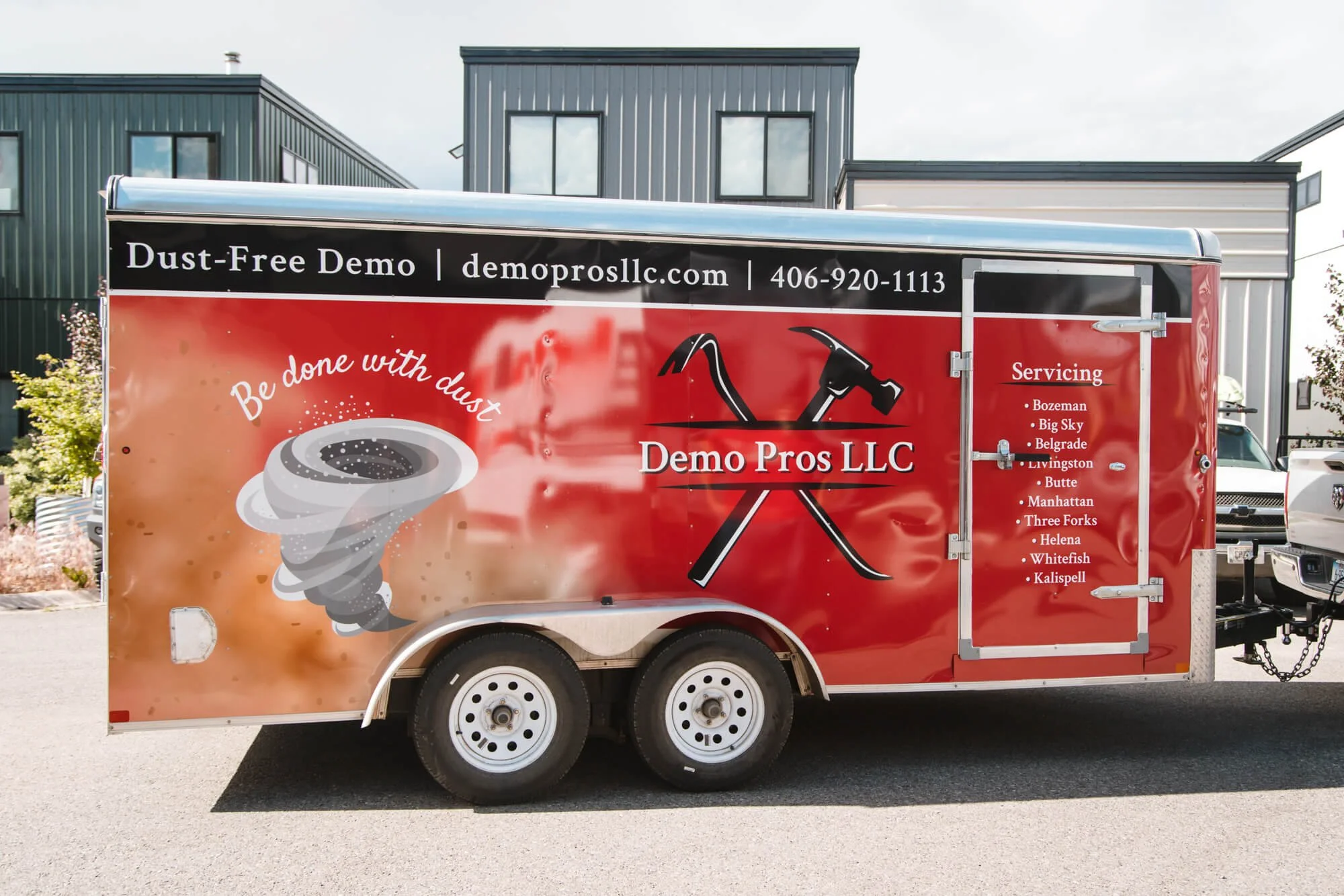 Side view of Demo Pros LLC trailer in Bozeman, Montana, with bold red trailer graphics featuring tools, a dust funnel illustration, tagline 'Be done with dust,' and a service list of Montana locations. 