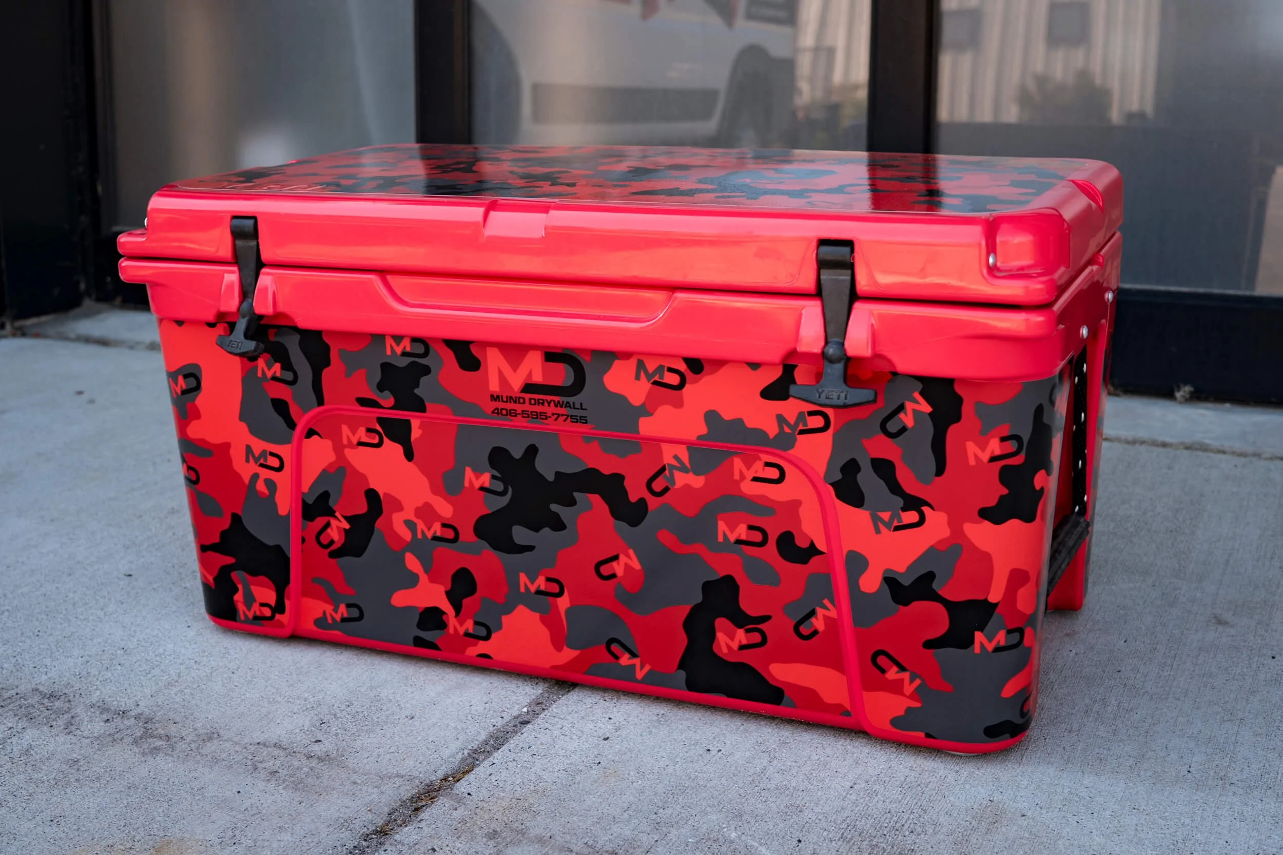 Bright red YETI 65 with a full vinyl wrap on the sides and lid. Red and camo pattern with Mund Drywall logo scatter throughout