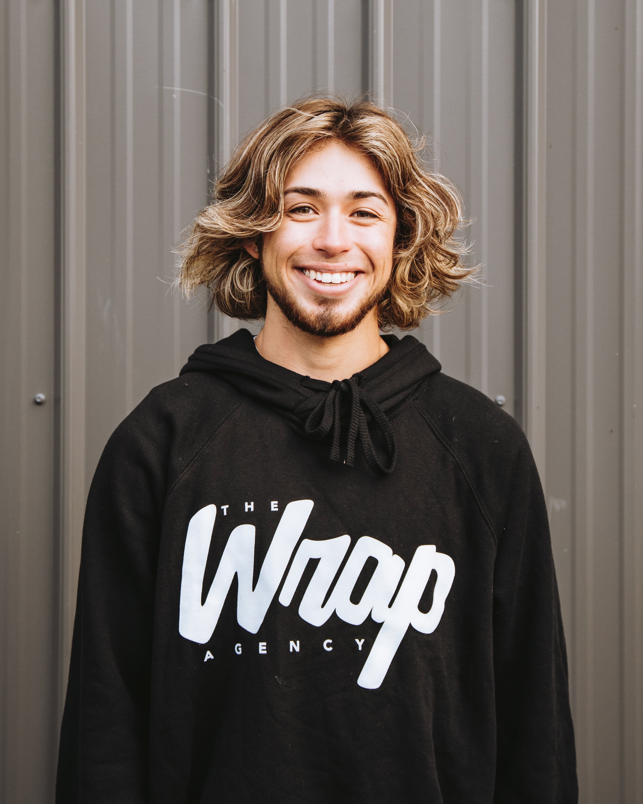 Person with shoulder-length hair smiling, wearing a black hoodie with "The Wrap Agency" logo, standing in front of a gray corrugated metal wall.