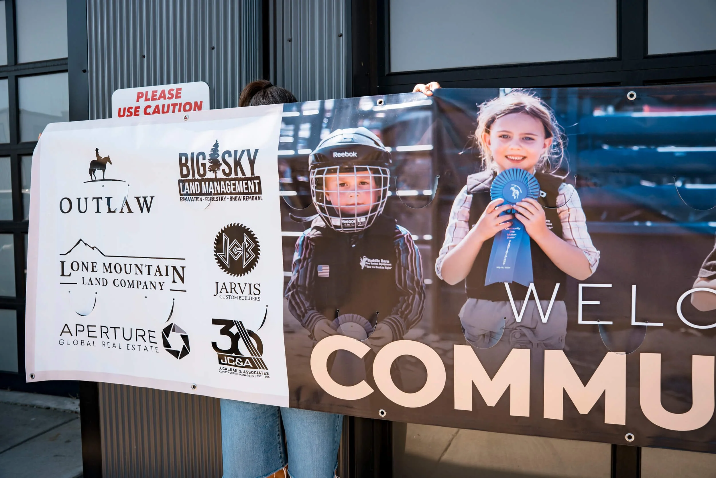 Closeup of an extra large banner featuring a photo of Mutton Busting kids and sponsor logos, promoting Big Sky PBR's Community Day. Visible wind slits