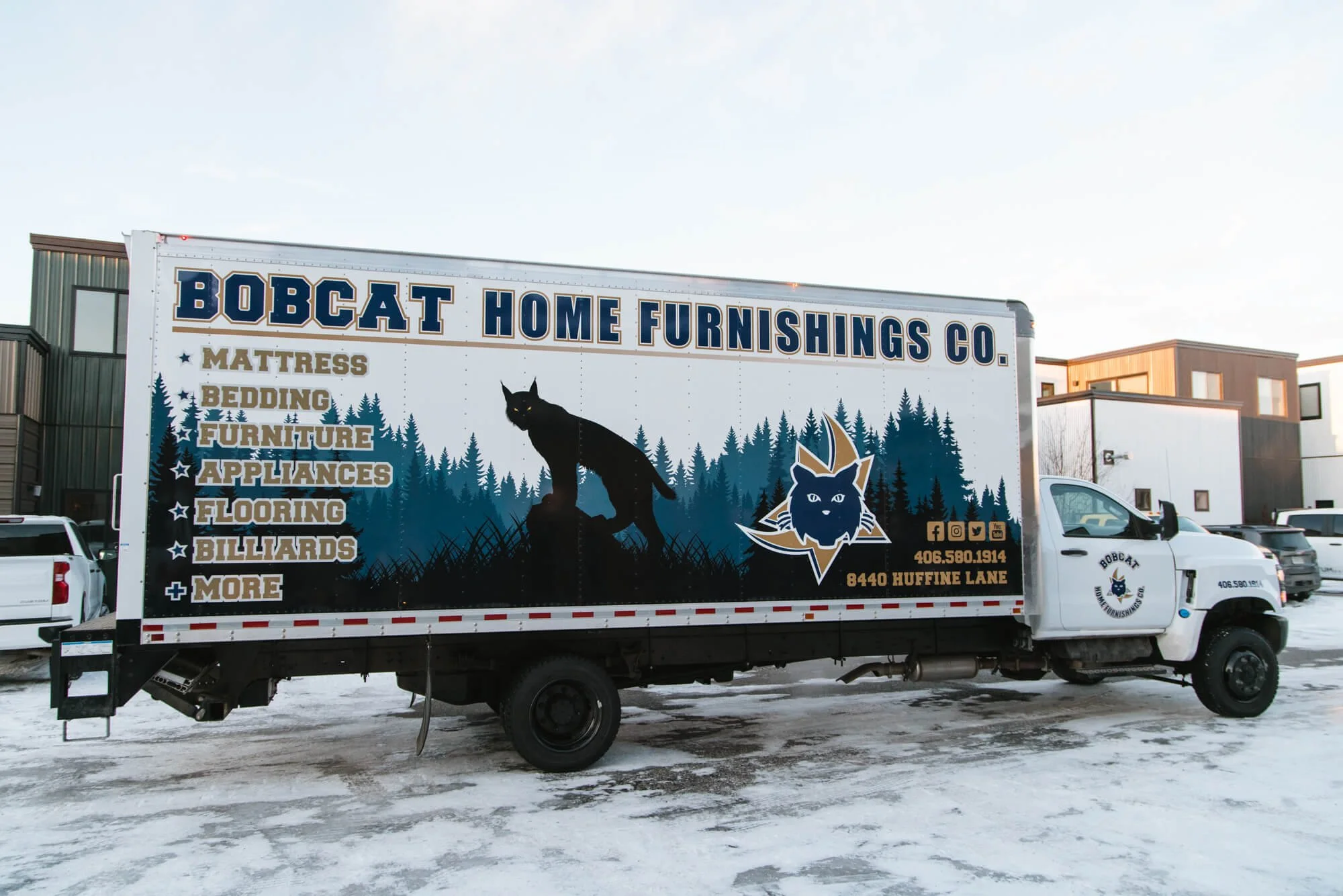 Large box truck with a bold vehicle wrap for Bobcat Home Furnishings Co. in Bozeman, MT, featuring a mountain forest graphic, a silhouetted bobcat, and a full list of home products like furniture, bedding, and appliances.