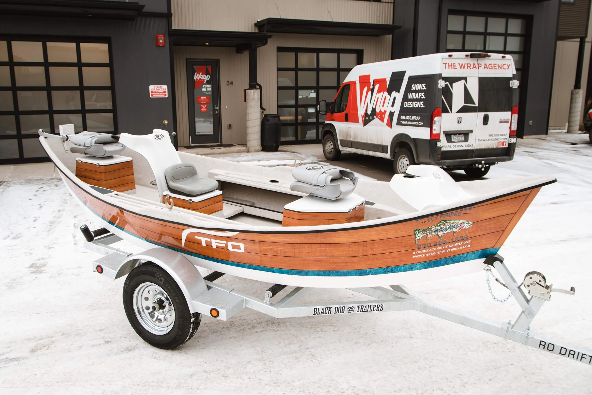 Custom vinyl wrap applied to a white drift boat with woodgrain-effect sides and teal detailing, parked in front of The Wrap Agency van and garage in Bozeman, MT.