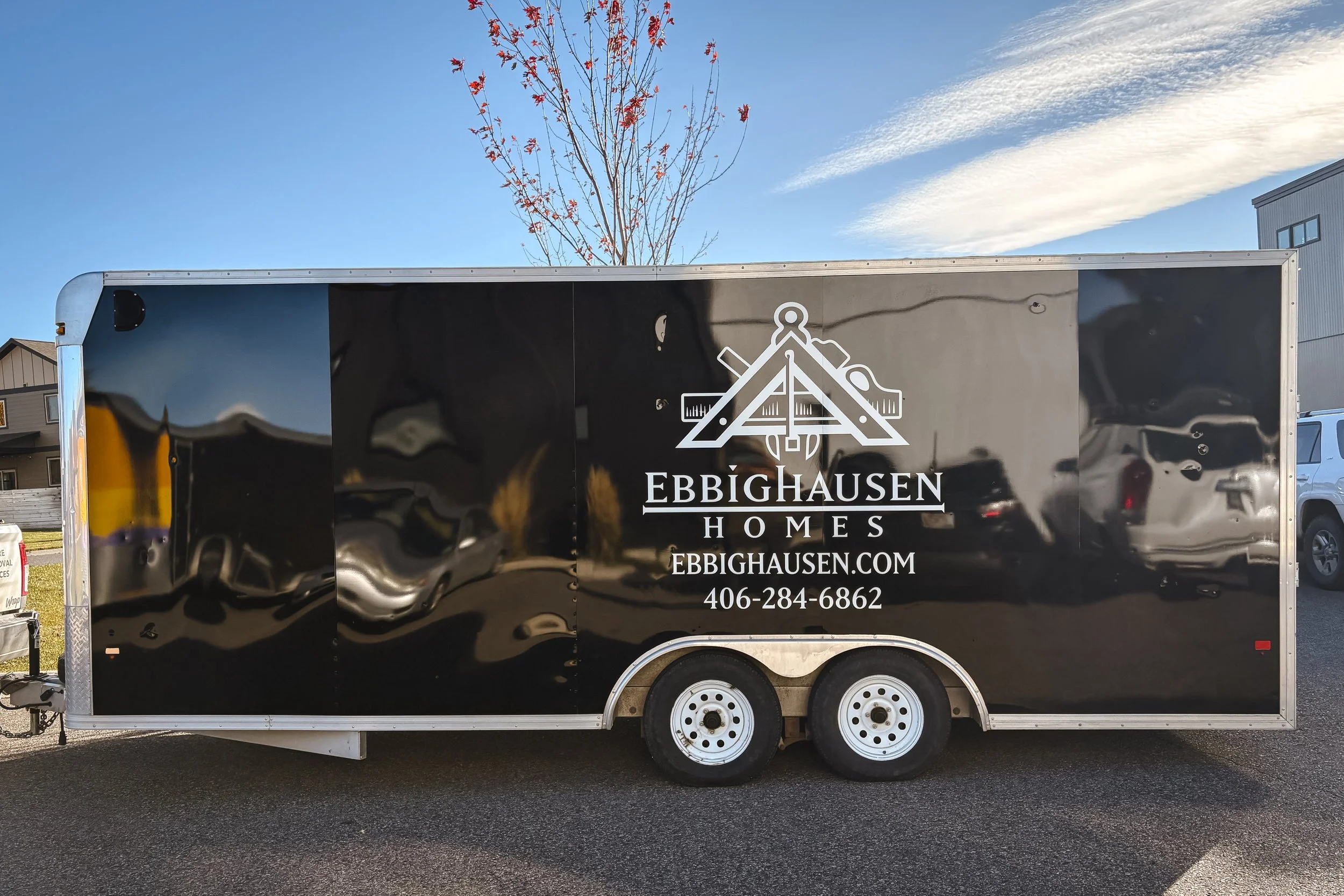 Side view of a black trailer with a white Ebbighausen Homes logo centered over the wheel well. Their contact info is centered beneath the logo