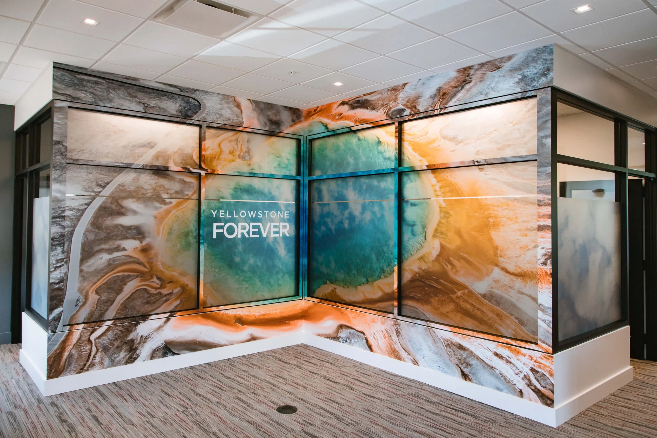 Floor-to-ceiling wall wrap displaying a vibrant aerial image of Yellowstone’s Grand Prismatic Spring with the words “Yellowstone Forever” centered on the glass-paneled wall. Business Location: Yellowstone Forever in Bozeman, MT. 