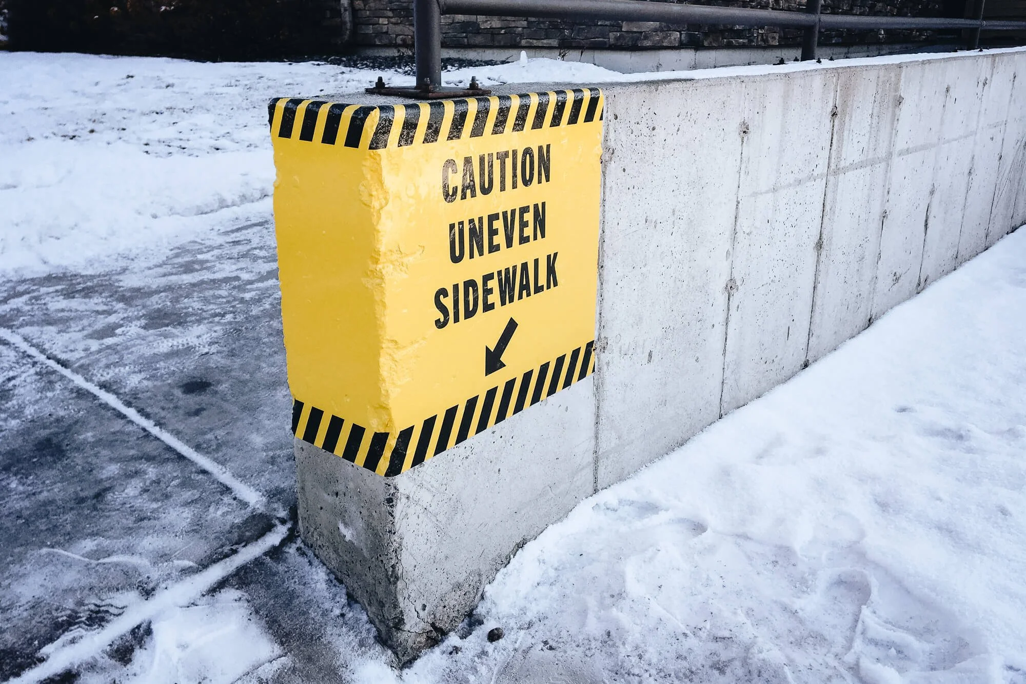 Bright yellow “Caution Uneven Sidewalk” warning printed as a custom vinyl wrap applied to a concrete pillar to enhance pedestrian safety in winter conditions. Custom vinyl wraps for outdoor safety messaging.