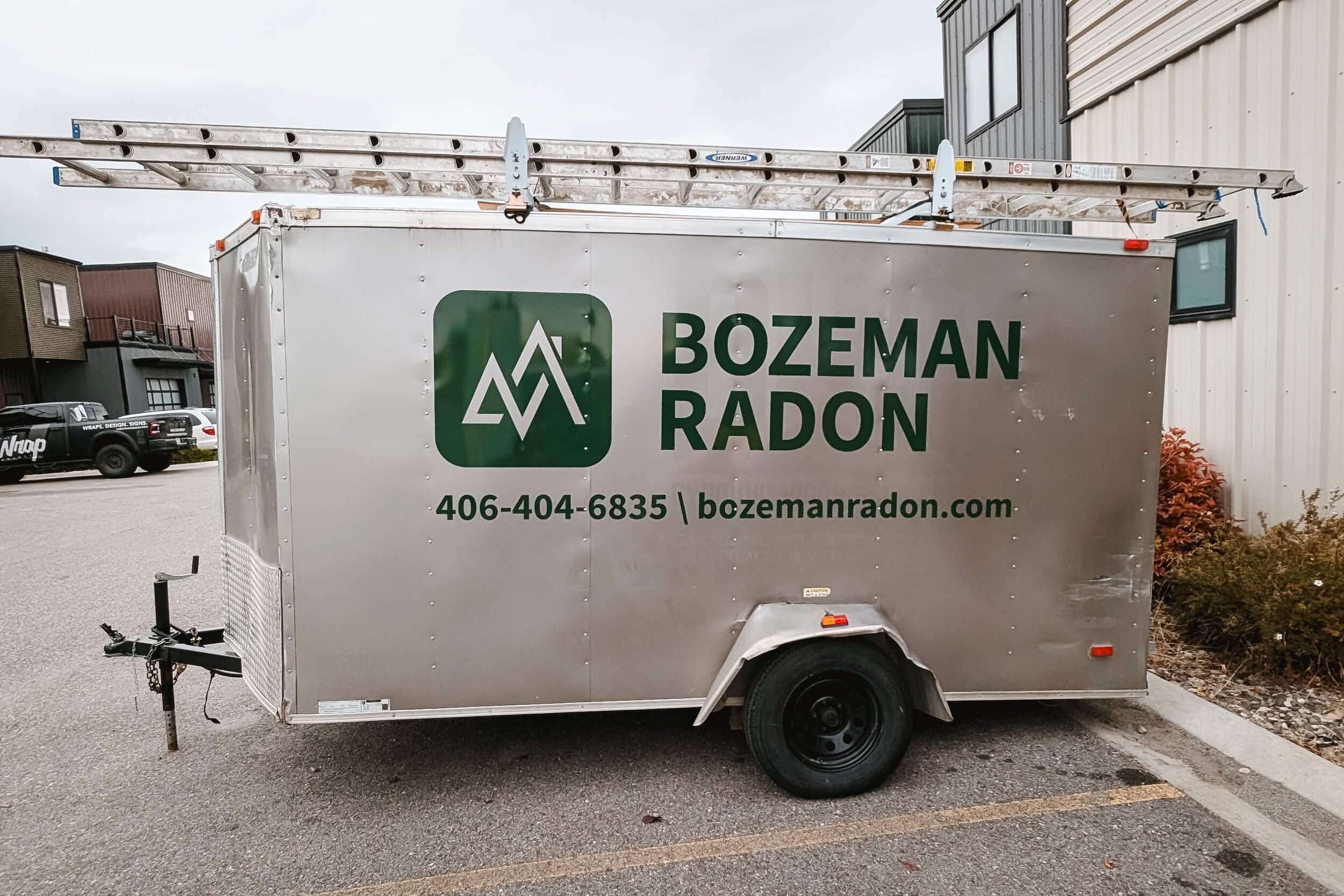 Driver-side view of a silver trailer with a large vinyl logo for Bozeman Radon with contact info below