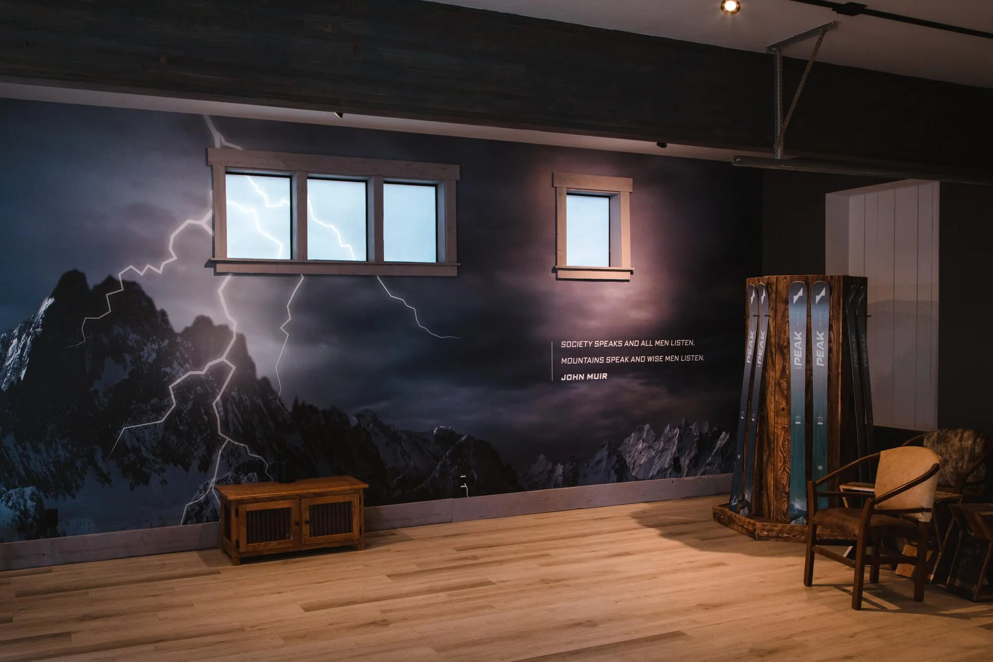 Indoor space with a mountain-themed wall wrap featuring lightning and a John Muir quote, wooden floor, a small table, chairs, and a stand with skis.