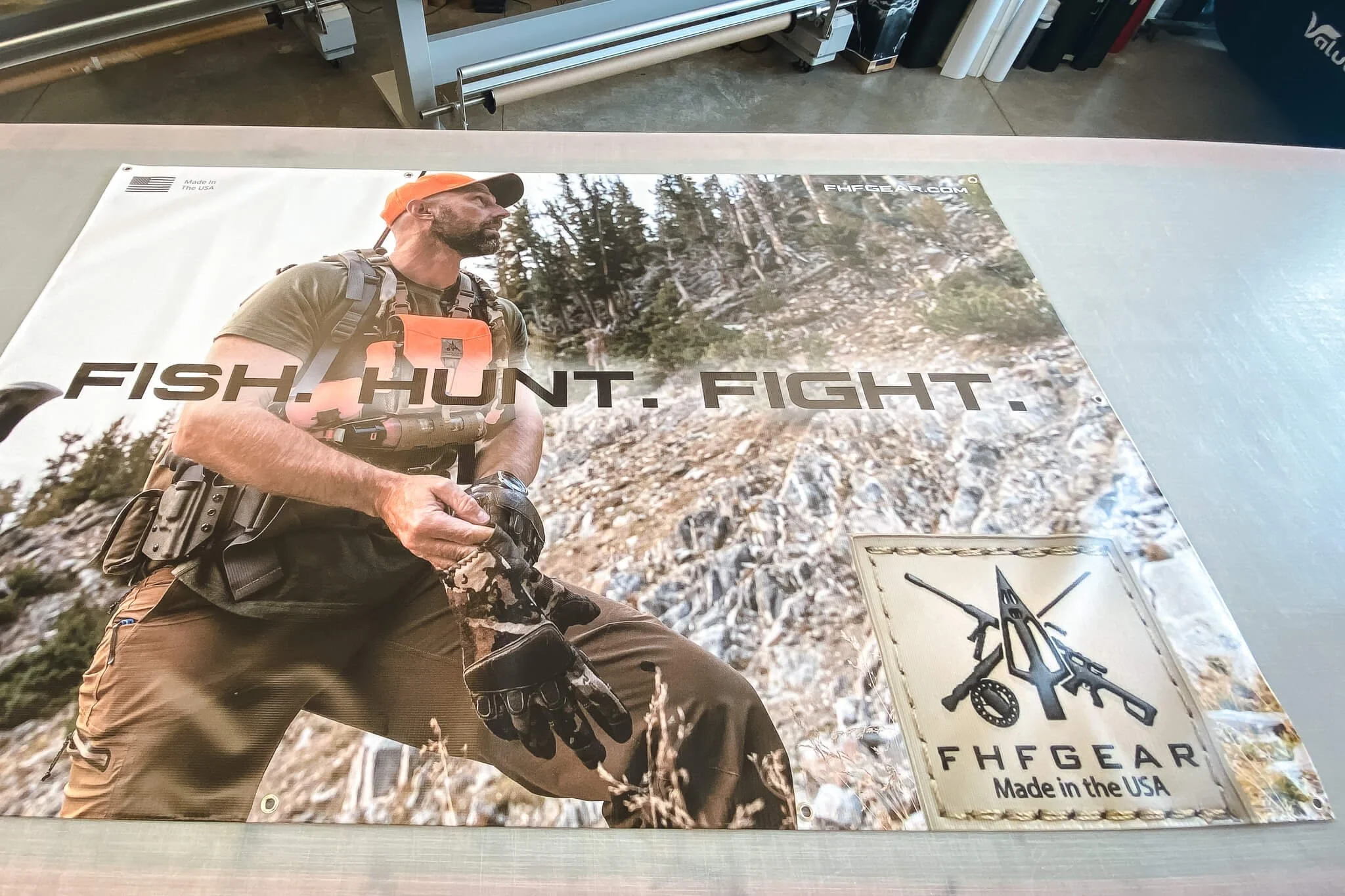 High-resolution vinyl banner printing for FHF Gear displaying the phrase 'Fish. Hunt. Fight.' with a close-up of a rugged outdoorsman in full gear.